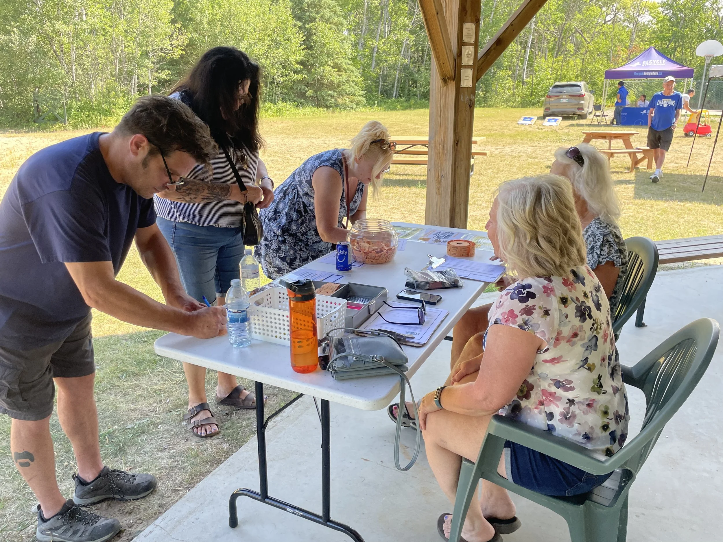 People sitting and standing around a registration or sign-in table outdoors under a wooden pavilion, with a grassy field and wooded area in the background. The table has papers, water bottles, and a plastic container with snacks.