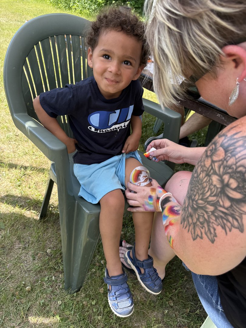 A young boy sitting in a green outdoor chair with a slightly confused expression, as a woman applies face paint to his leg during an outdoor event.