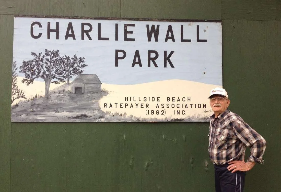 An elderly man wearing glasses, a white cap, and a plaid shirt stands next to a sign for Charlie Wall Park, Hillside Beach Ratepayer Association, 1982.