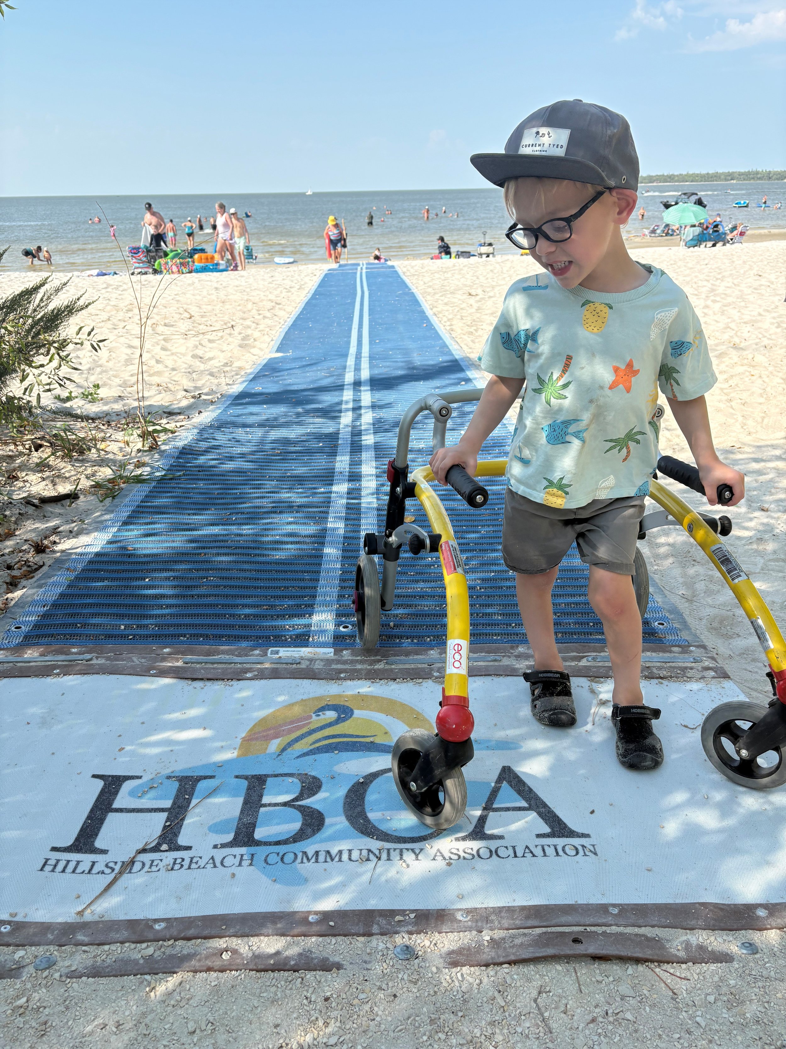 A young boy wearing a Hawaii-themed shirt, shorts, and a hockey cap with glasses, walking on a blue ramp at the beach with a walker, with people and the ocean in the background.