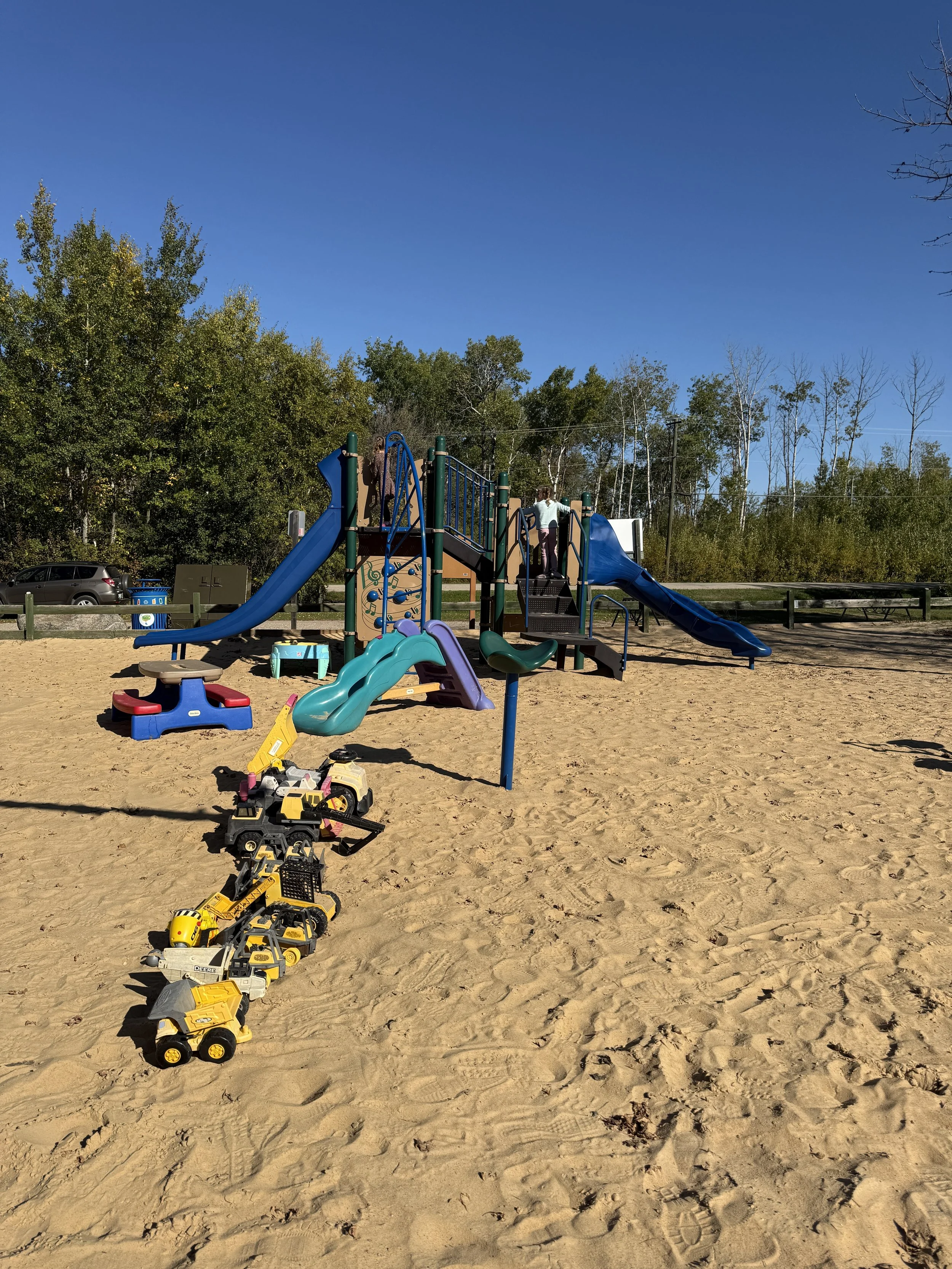 Children's playground with slides, climbing structures, and sand surface, with toy trucks in the foreground and trees in the background under a clear blue sky.