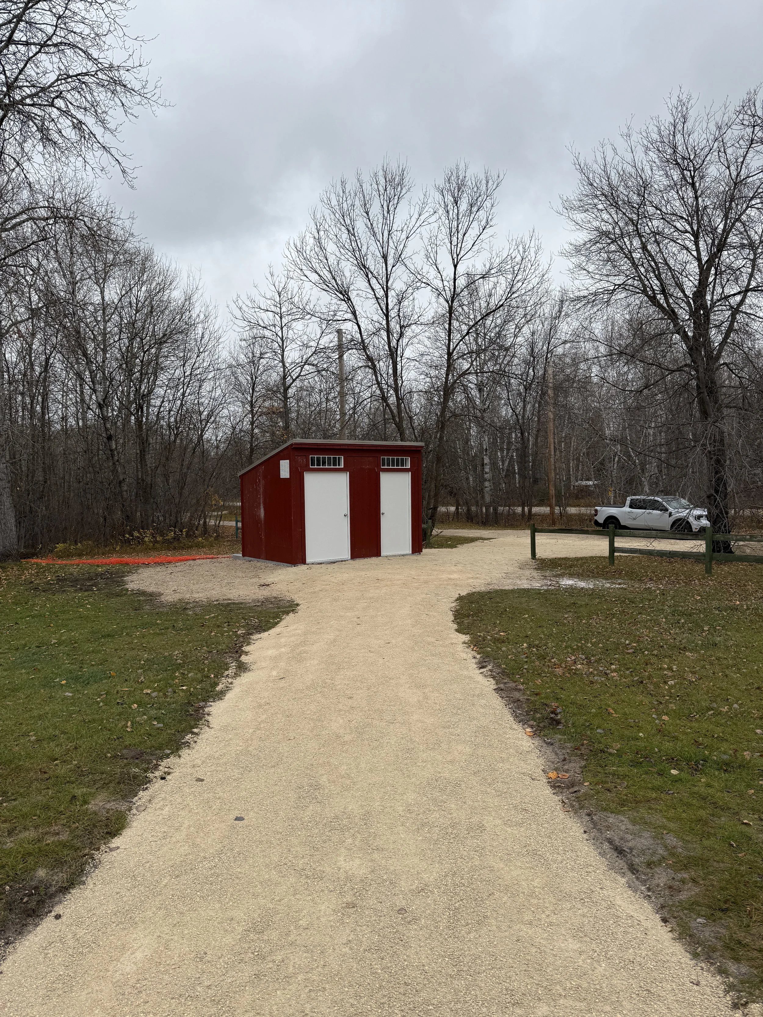 A gravel pathway leading to a red and white small building with two doors, surrounded by bare trees and a grassy area, on a cloudy day.
