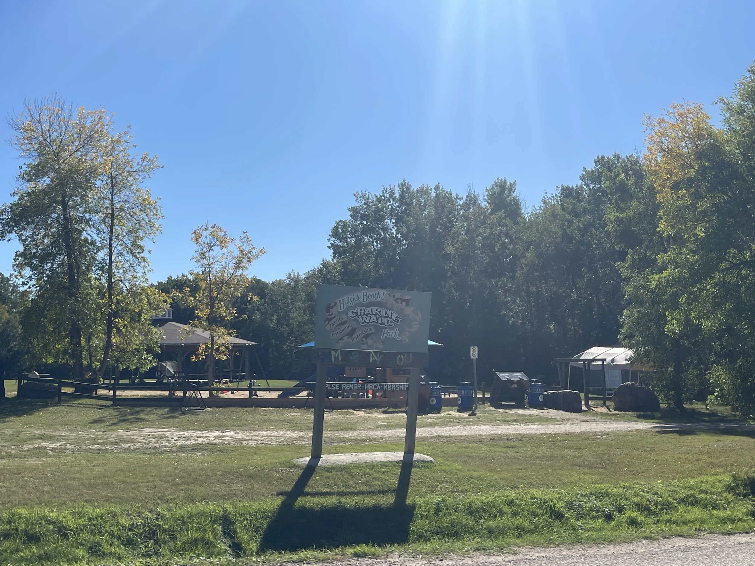 A signboard in front of Hillside Beach's Charlie Wall's Park with some trees, a building, and trash bins in the background under a clear blue sky.
