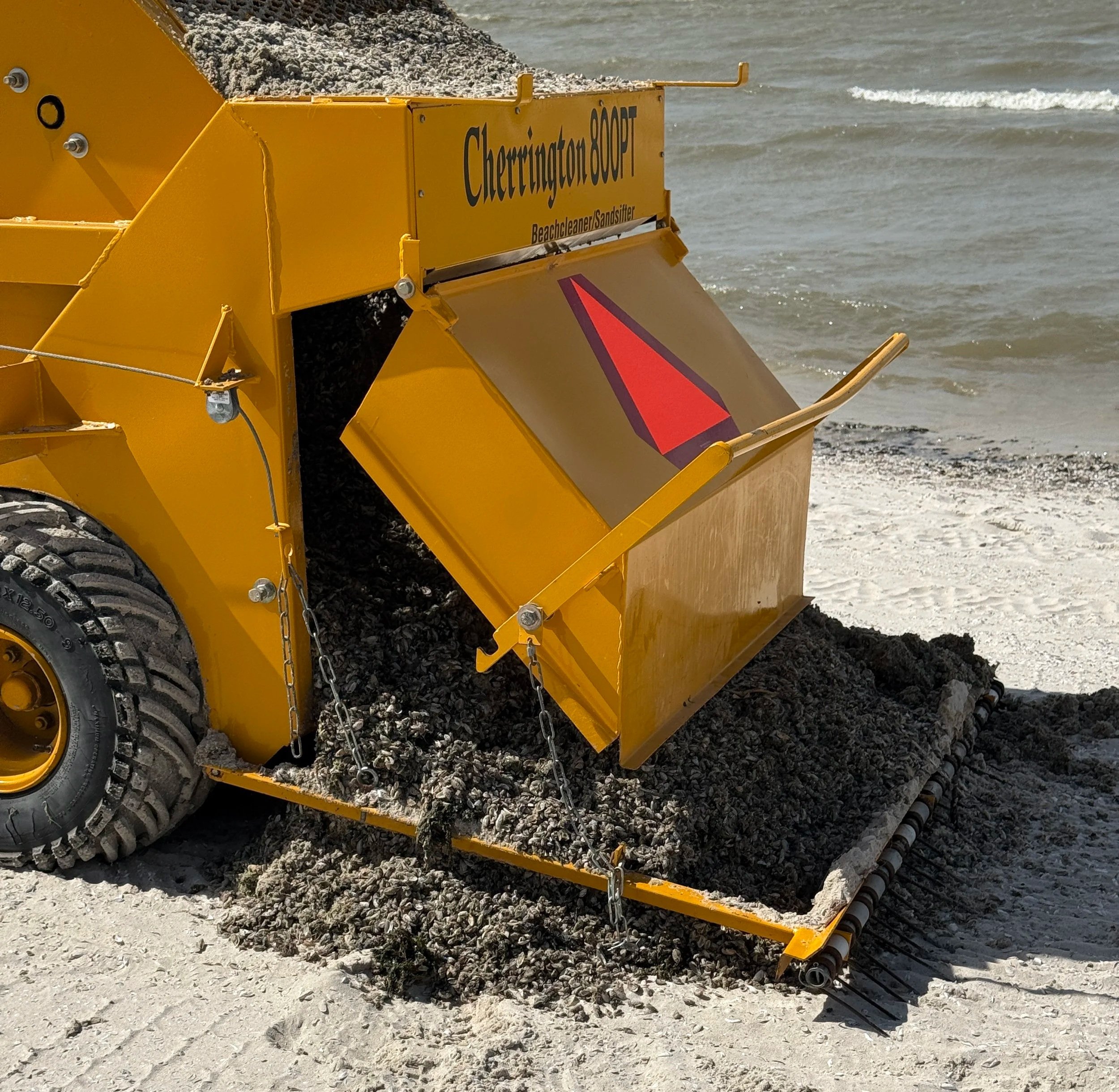 A yellow beach cleaning machine labeled "Cherrington 600PT" with a slide hopper collecting sand and debris at the beach shoreline with water in the background.