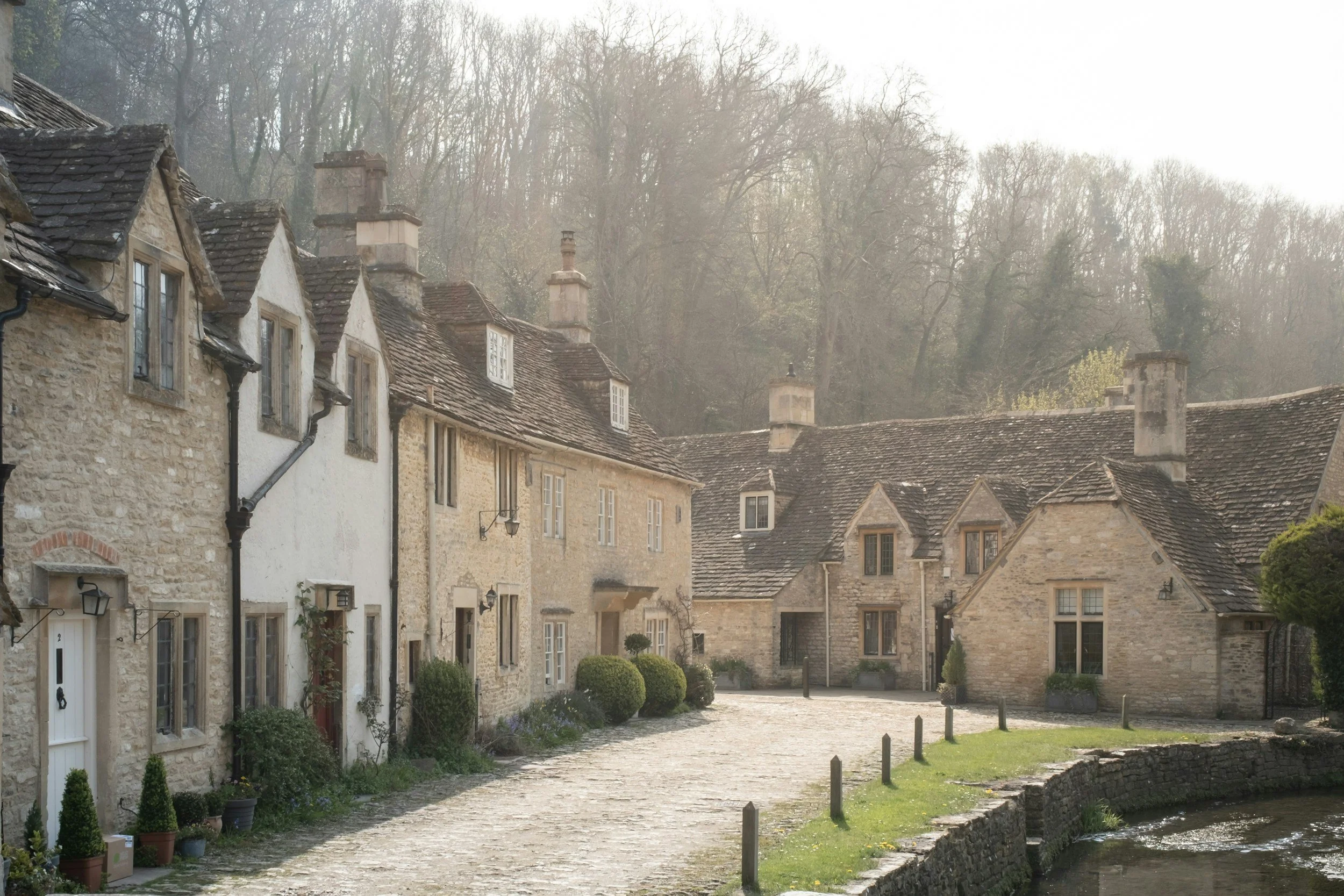 A row of historic stone cottages with sloped roofs and chimneys, along a cobblestone pathway beside a small stream.