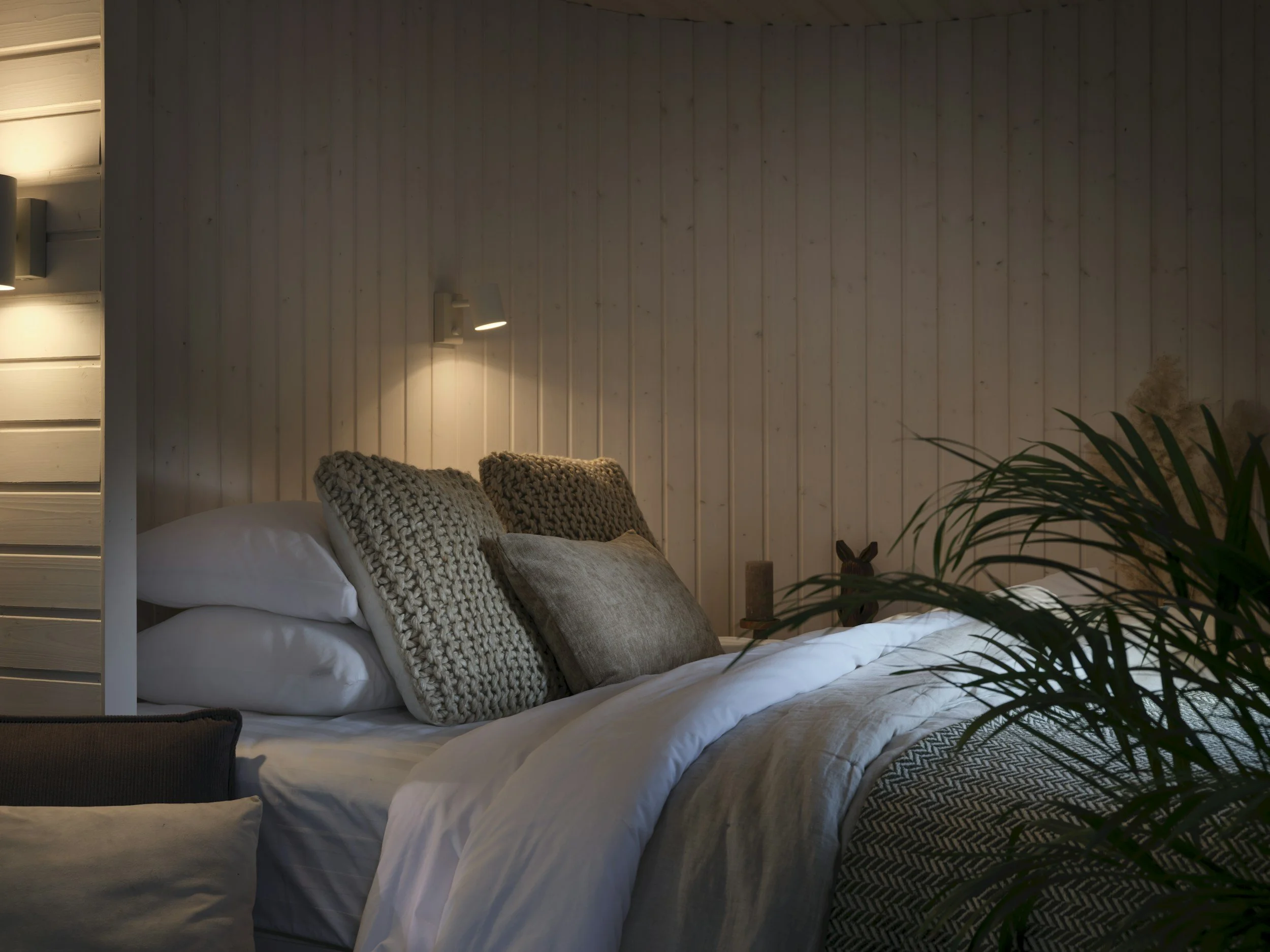 Cozy bedroom with neatly made bed, textured pillows, and warm lighting on a wooden wall backdrop, with a plant partially visible in the foreground.