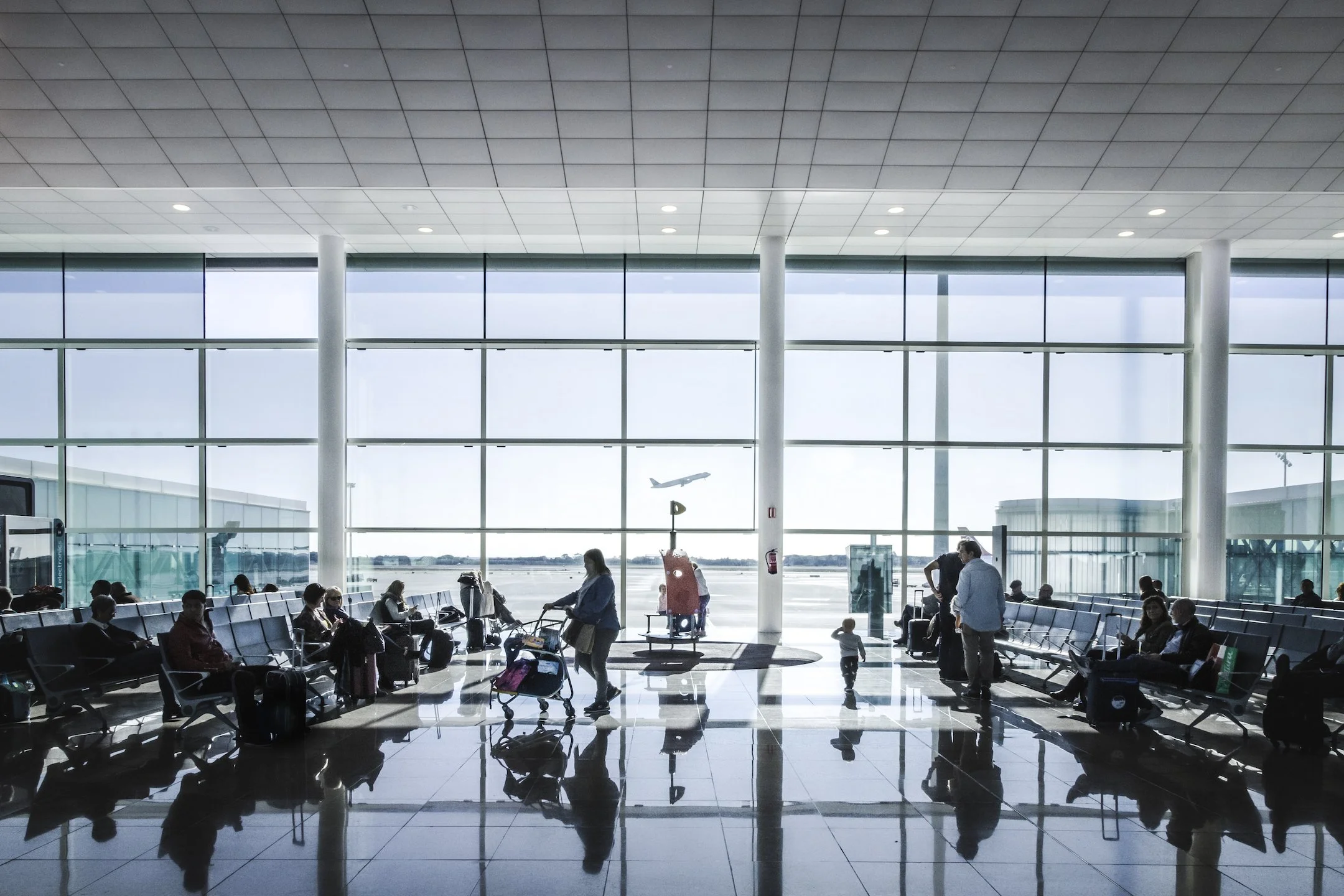 Salle d'attente dans un aéroport avec des passagers assis, des valises, et un avion en vol visible à travers les grandes fenêtres.