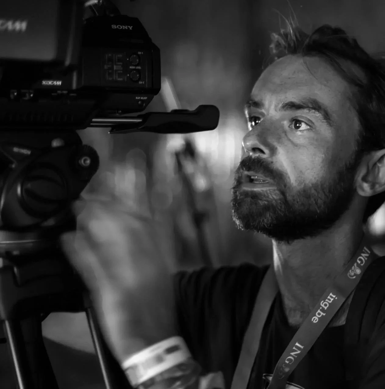Un homme avec une barbe et un tatouage, regarde à travers un viseur de caméra professionnelle lors du tournage, en noir et blanc.