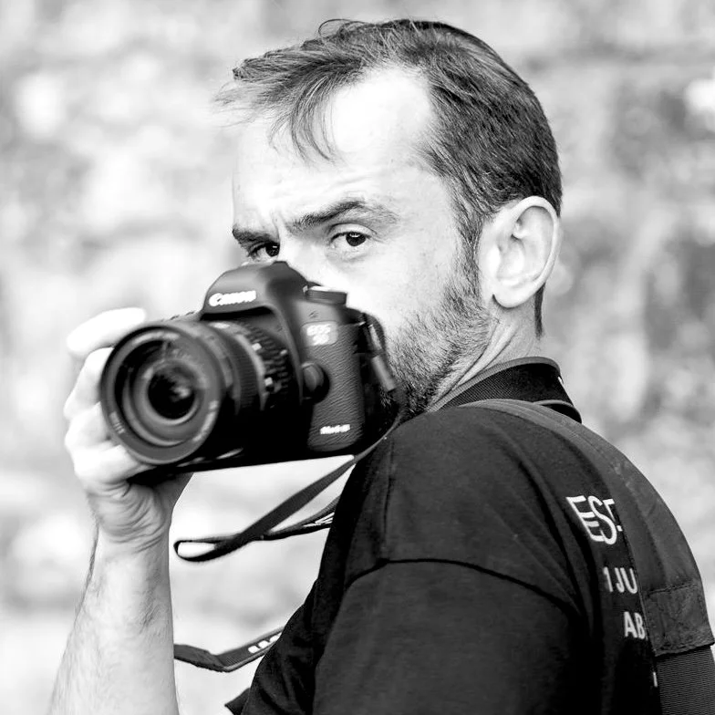 Un homme tient un appareil photo Canon en pointant vers l'avant, portant un T-shirt noir et un sac à dos, dans un contexte en extérieur.