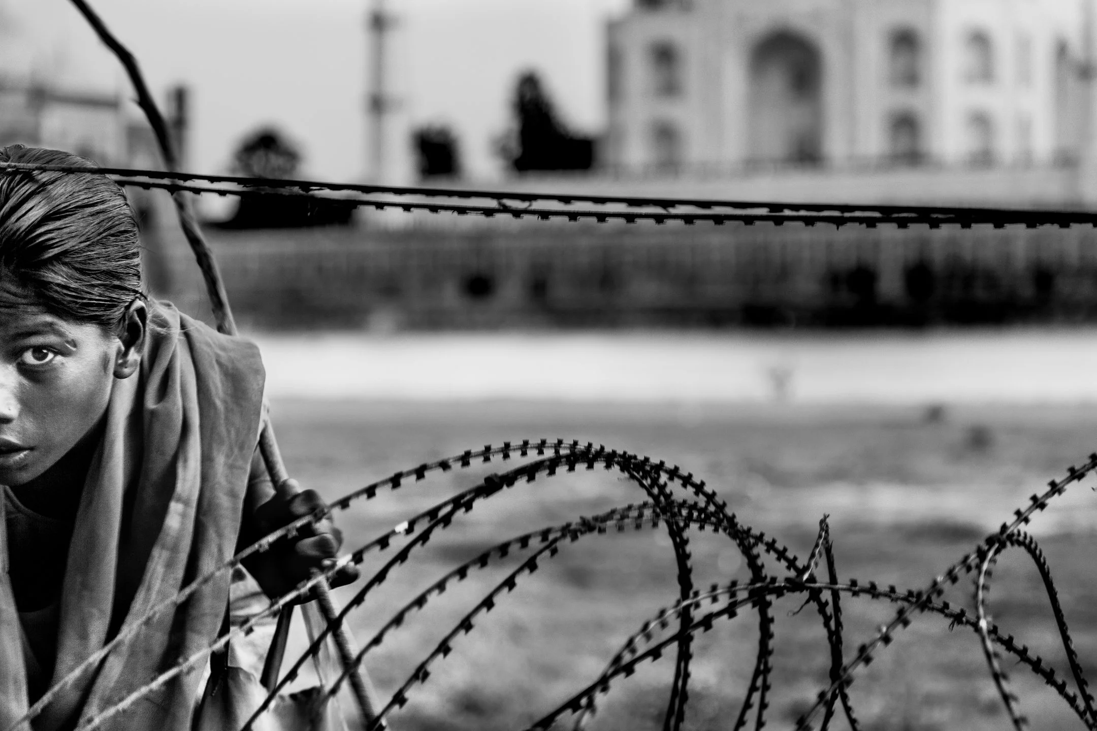 Personne tenant un fil barbelé, partialement visible, avec en arrière-plan des bâtiments flous.