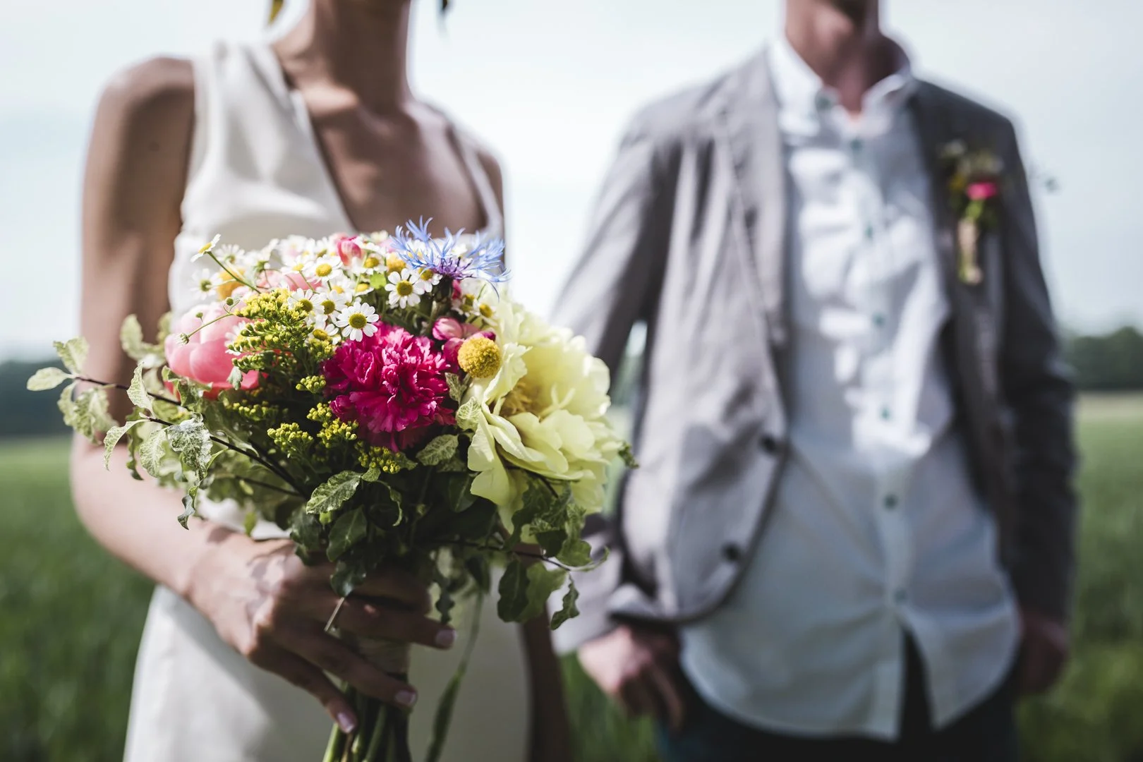 Une femme en robe blanche tient un bouquet de fleurs colorées lors d'un mariage en plein air, avec un homme à ses côtés dans un champ verdoyant.