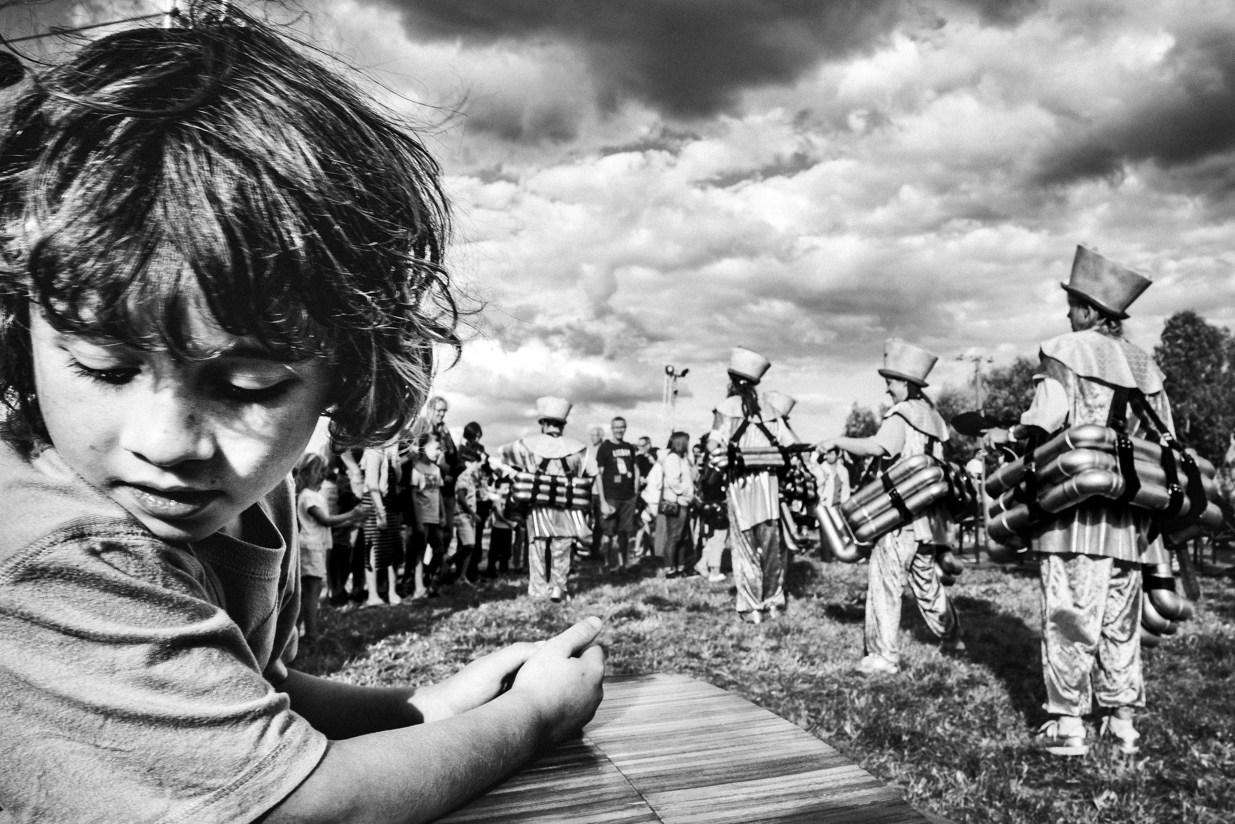Une jeune garçon regardant un spectacle de personnes déguisées en clowns ou personnages folkloriques avec des barils attachés, lors d'une fête en plein air avec un groupe de spectateurs et un ciel nuageux.