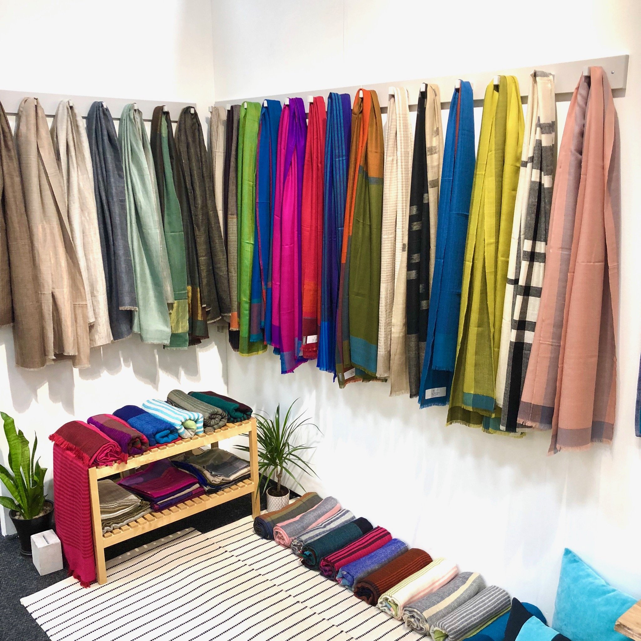 Display of colorful pashmina scarves hanging on a wall, with additional folded scarves on a wooden shelf and floor, and some potted plants in a retail setting in Shoppe Object Trade Show in New York.
