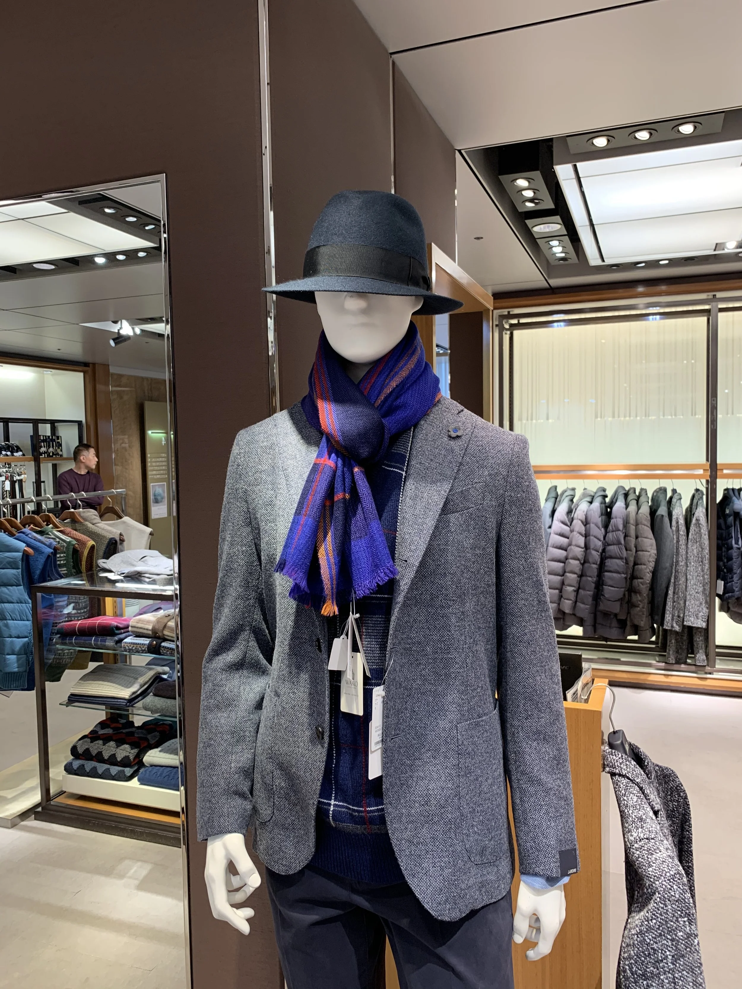 Mannequin dressed in a gray blazer, dark pants, with a blue and red plaid scarf, wearing a gray fedora hat with a black band, in a clothing store.