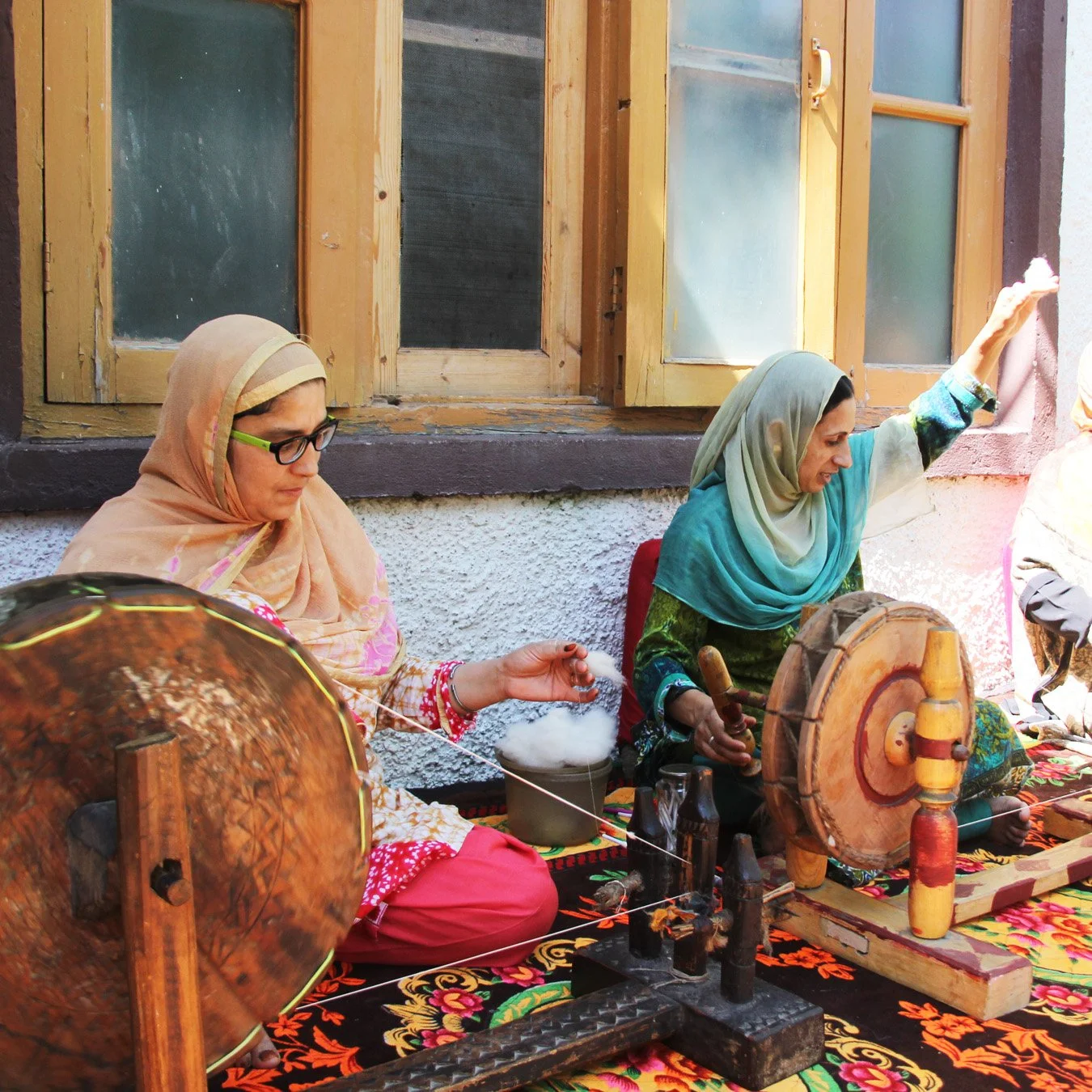 Two Kashmiri women wearing traditional headscarves sit on a colorful patterned cloth, operating vintage wooden spinning wheels to spin pashmina yarn outdoors near a building with yellow window frames in Srinagar, Kashmir.