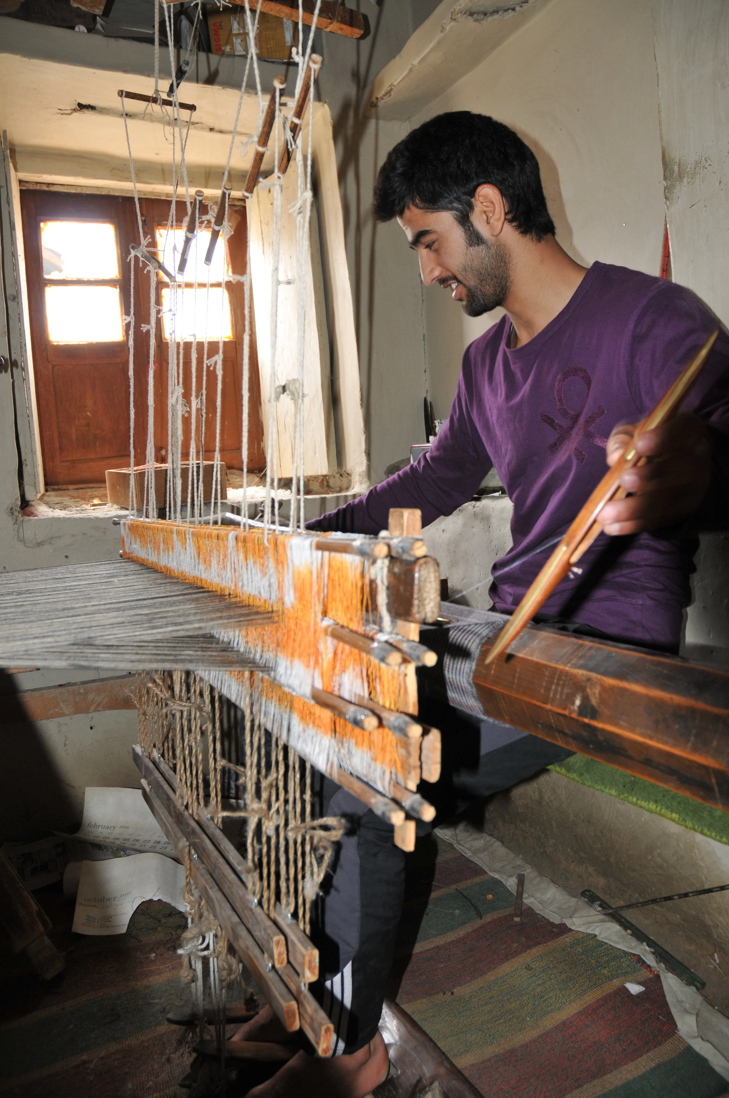 A man weaving on a traditional loom in a rustic room, with sunlight coming through a small window.