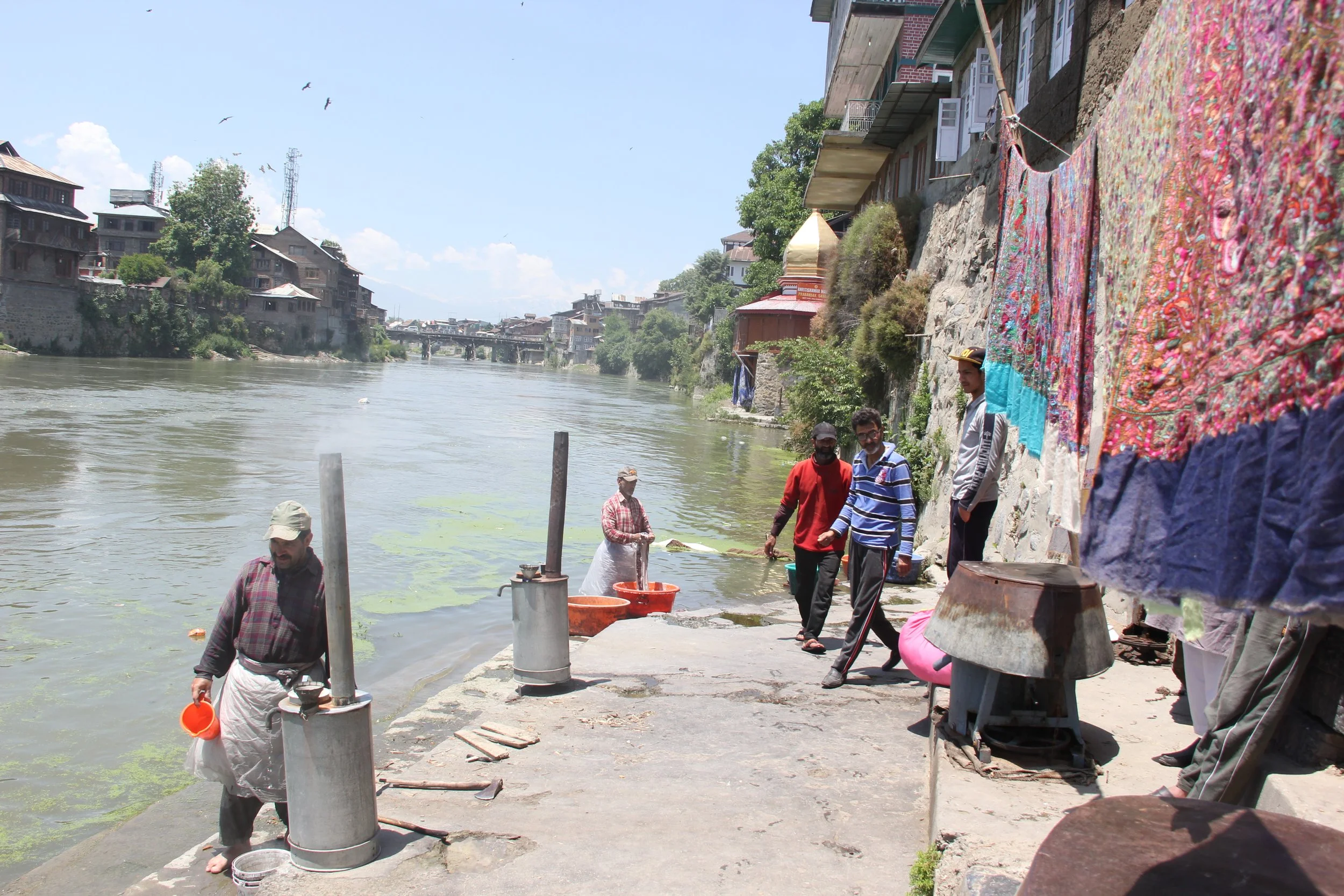 People gathering by a river, some washing clothes in the water, with residential buildings on a rocky riverbank and laundry hanging on a line, under a clear sky.