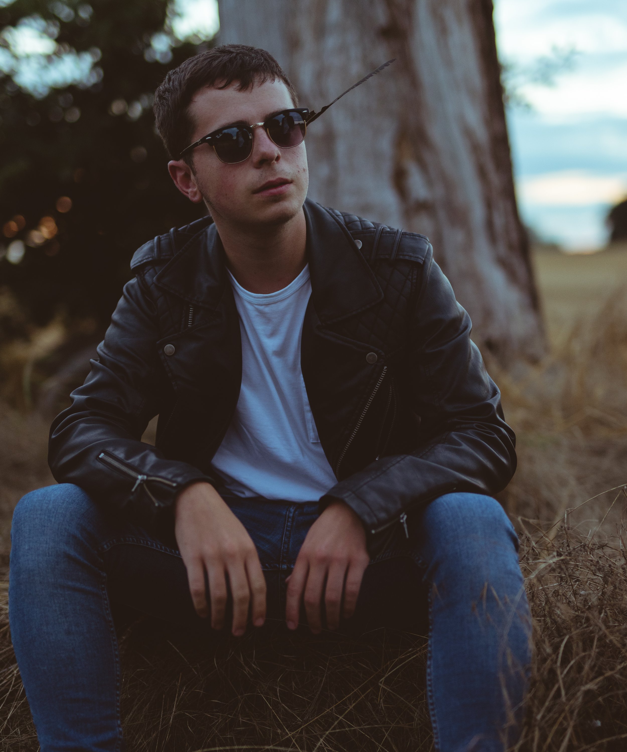 A young man with short dark hair, wearing sunglasses, a black leather jacket, a white shirt, and blue jeans, sitting outdoors on grass near a tree during sunset or late afternoon.