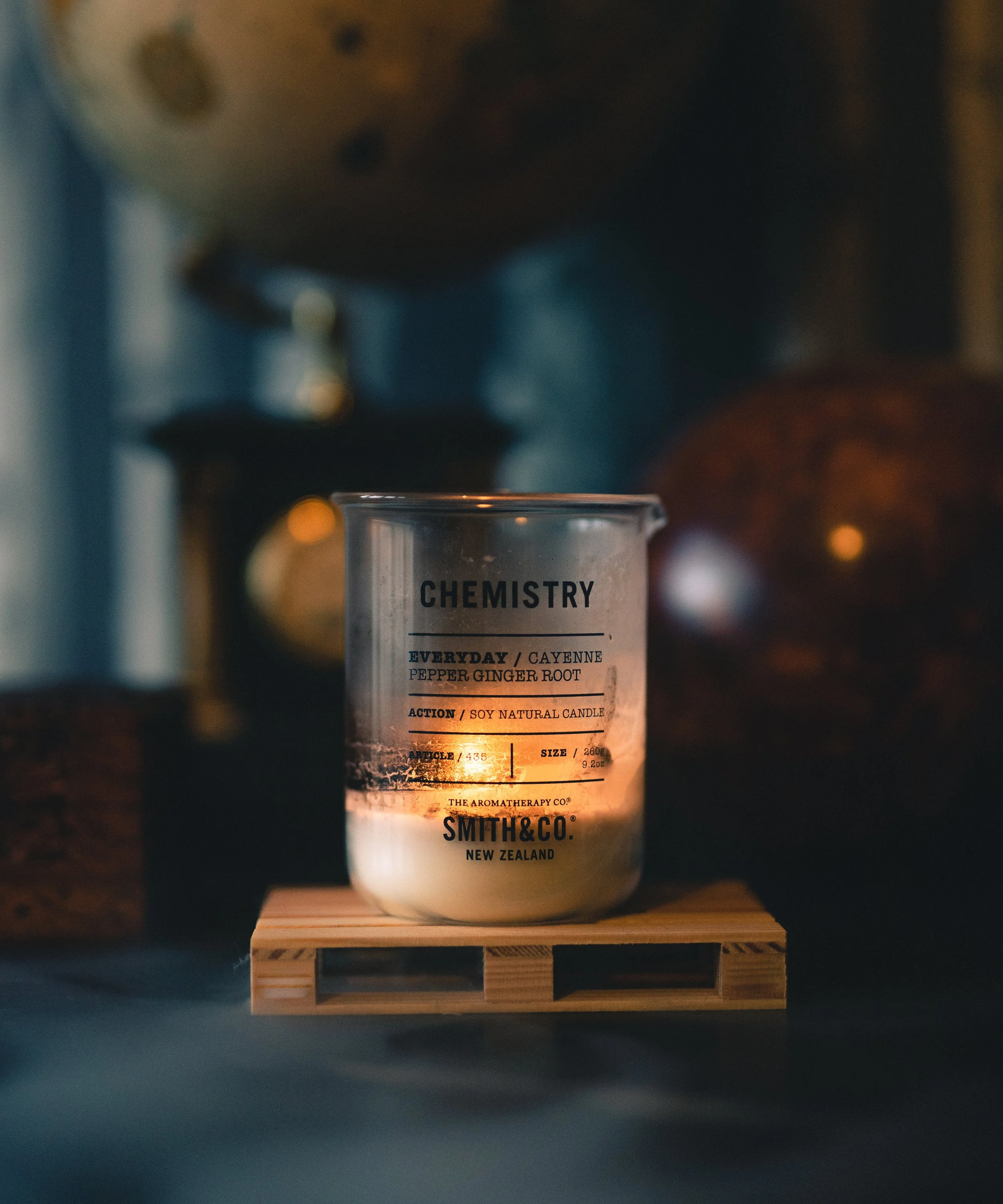A lit candle in a glass container labeled 'Chemistry' by Smith & Co. on a wooden surface, with a blurred background.
