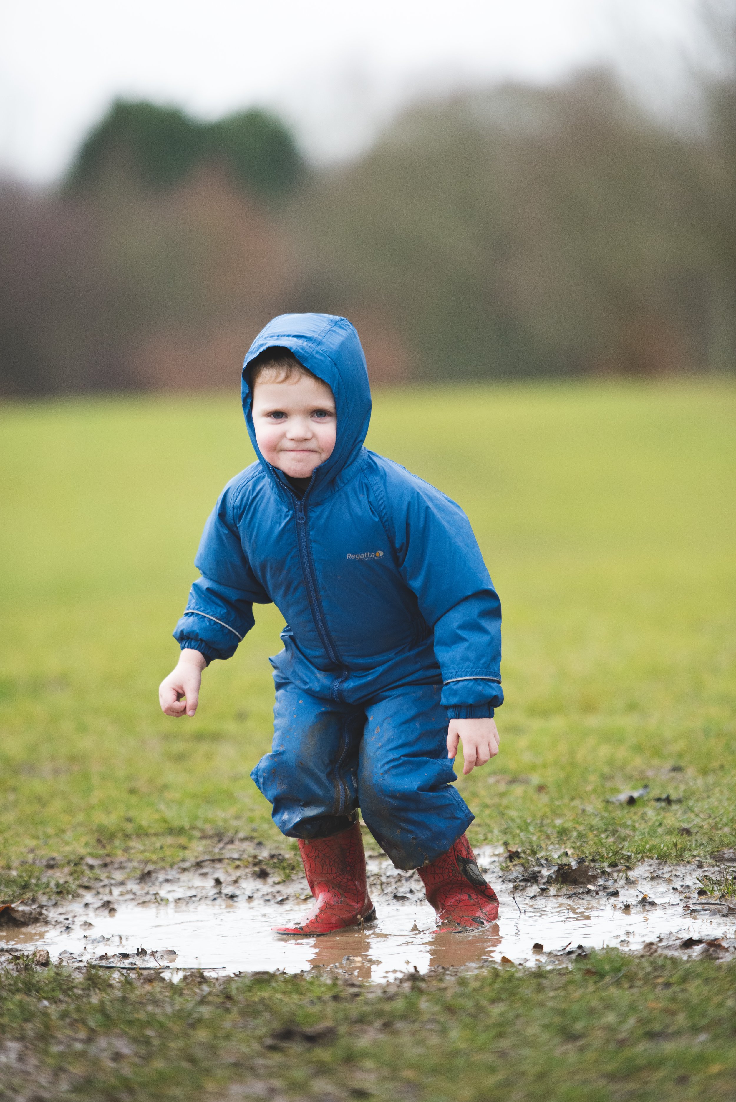 Young boy in a blue raincoat and red rain boots playing in a muddy puddle outdoors.