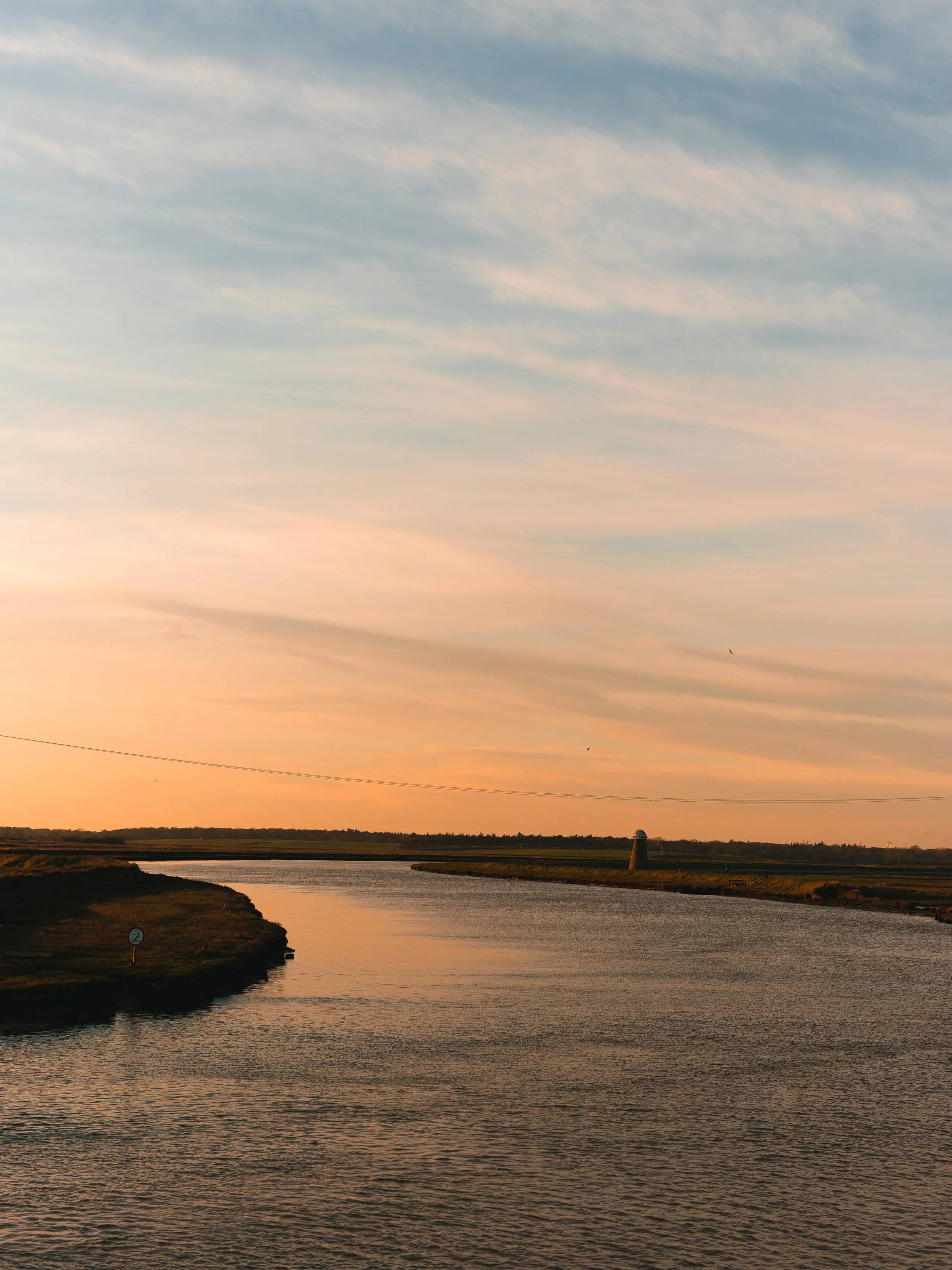 A river at sunset with a distant lighthouse, grassy banks, and a sky with wispy clouds.