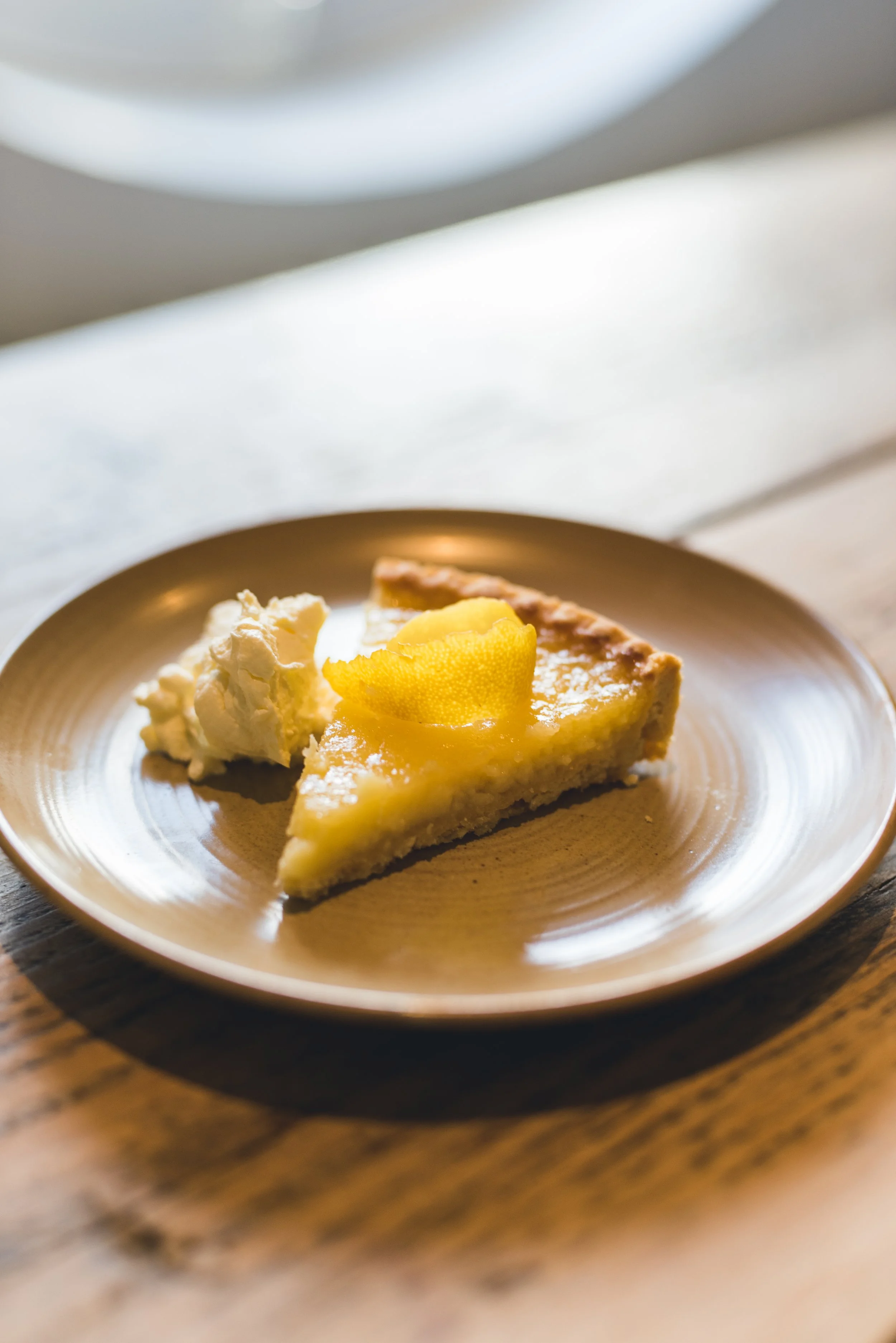 A slice of lemon pie with a dollop of whipped cream on a beige plate, placed on a wooden surface.