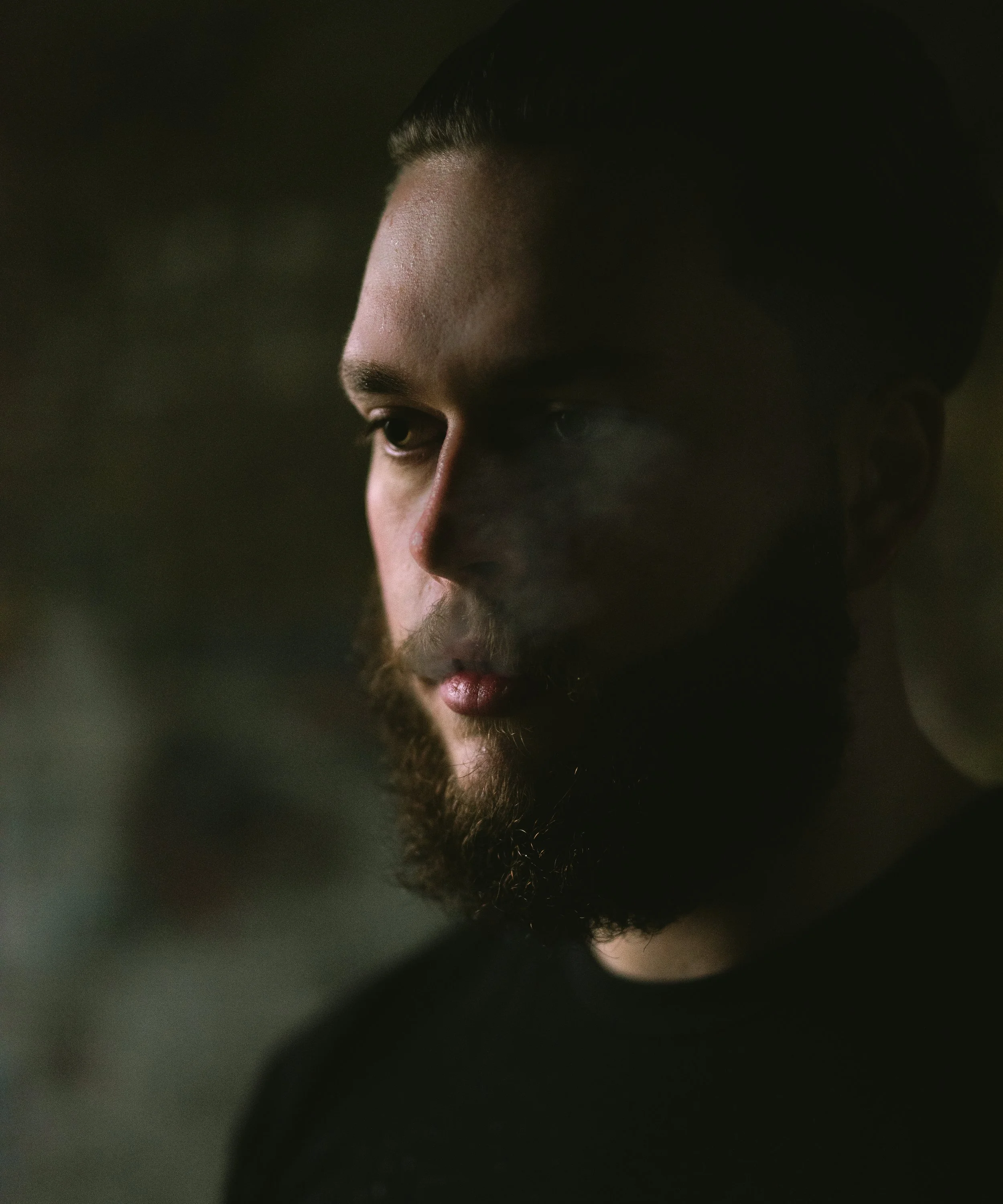Close-up of a man with a beard and slicked-back hair looking downward, with dark background and soft lighting.