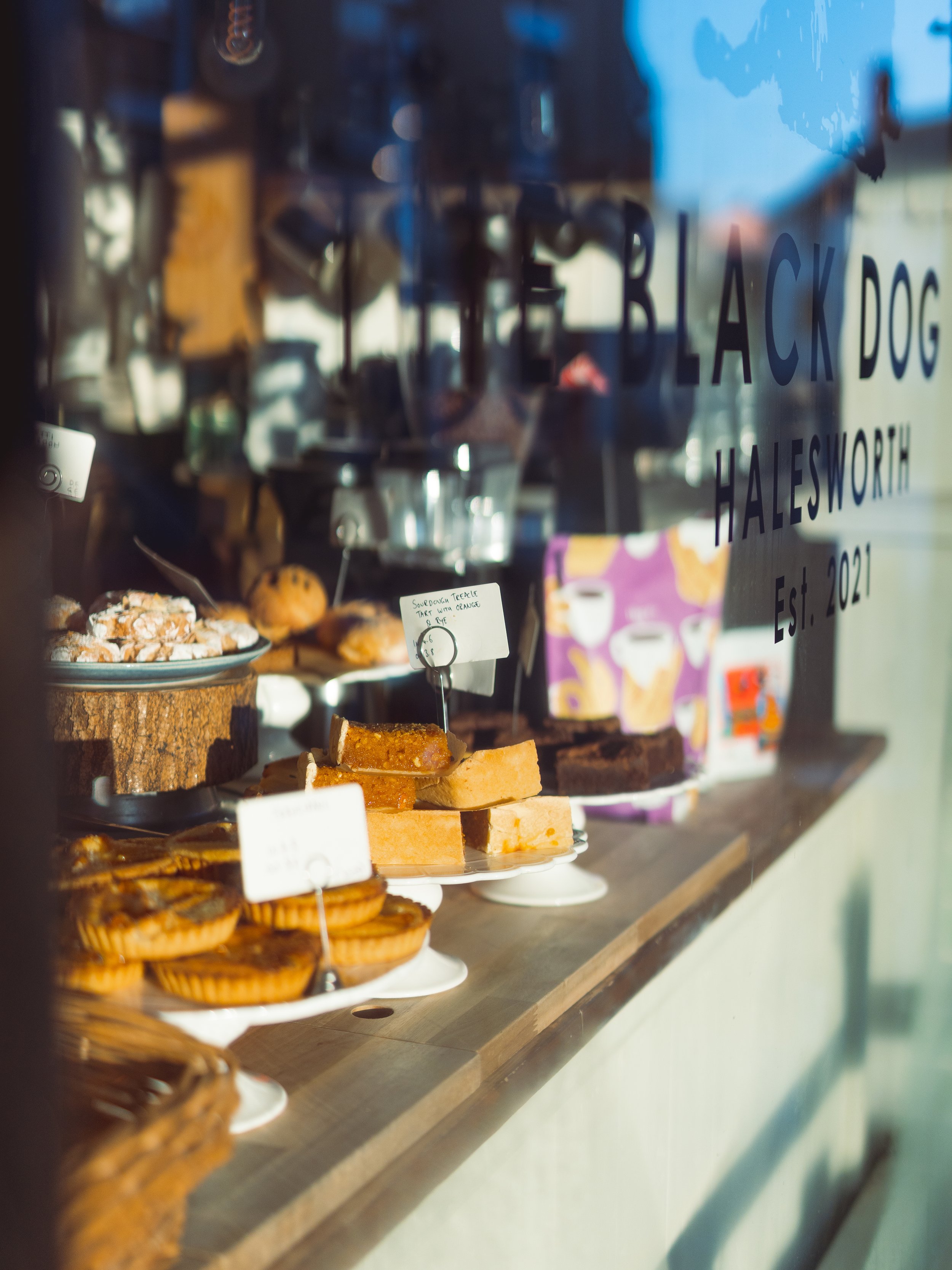 Display of various baked goods on a countertop in a shop window, including cookies, brownies, and bars, with the shop name 'Black Dog Halesworth' and establishment date '2021' written on the window.