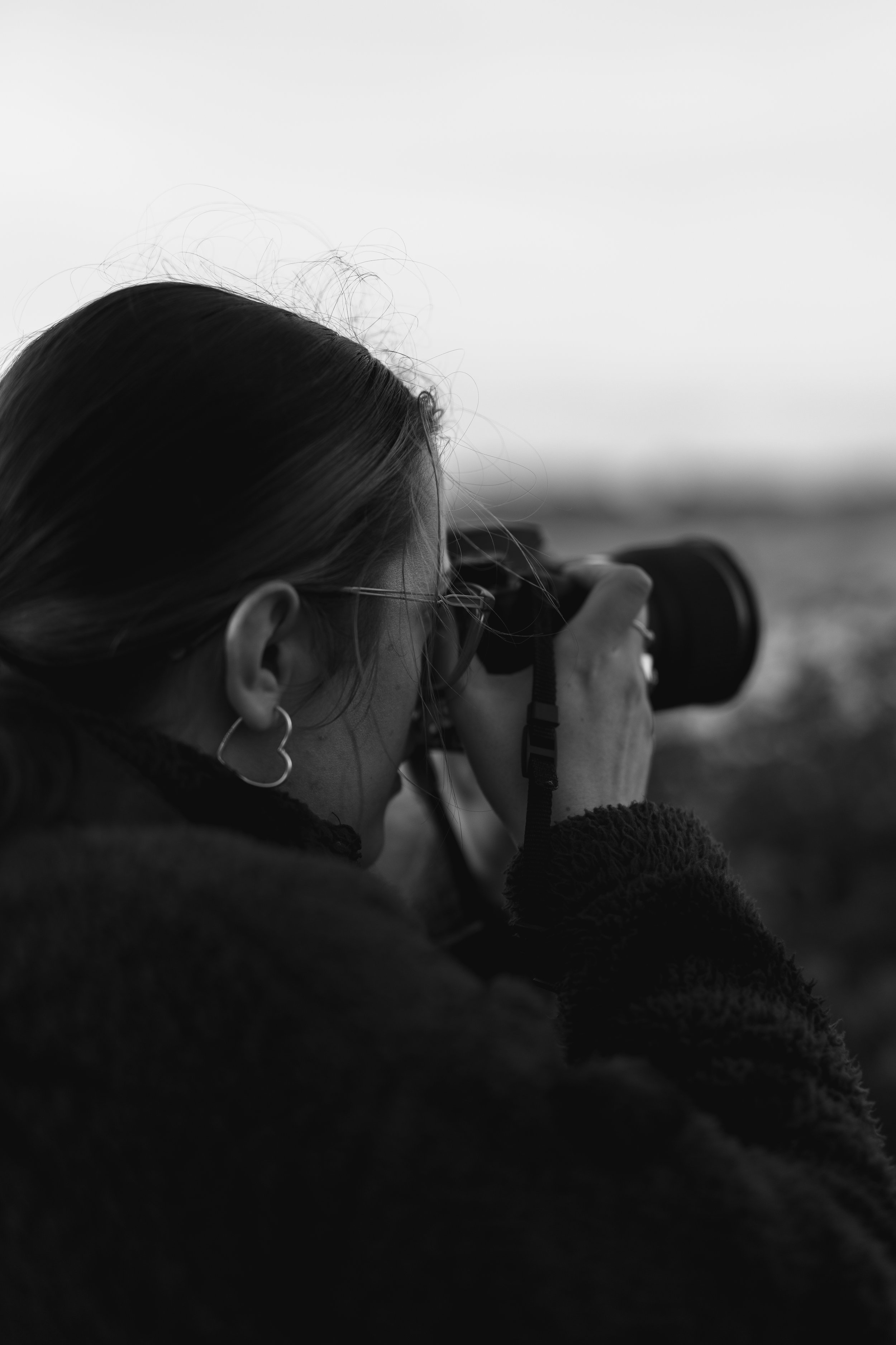 A woman with glasses and hoop earrings is taking a photograph with a camera. She has long hair and is wearing a dark fuzzy sweater. She is outdoors, and the background appears to be a blurred landscape.