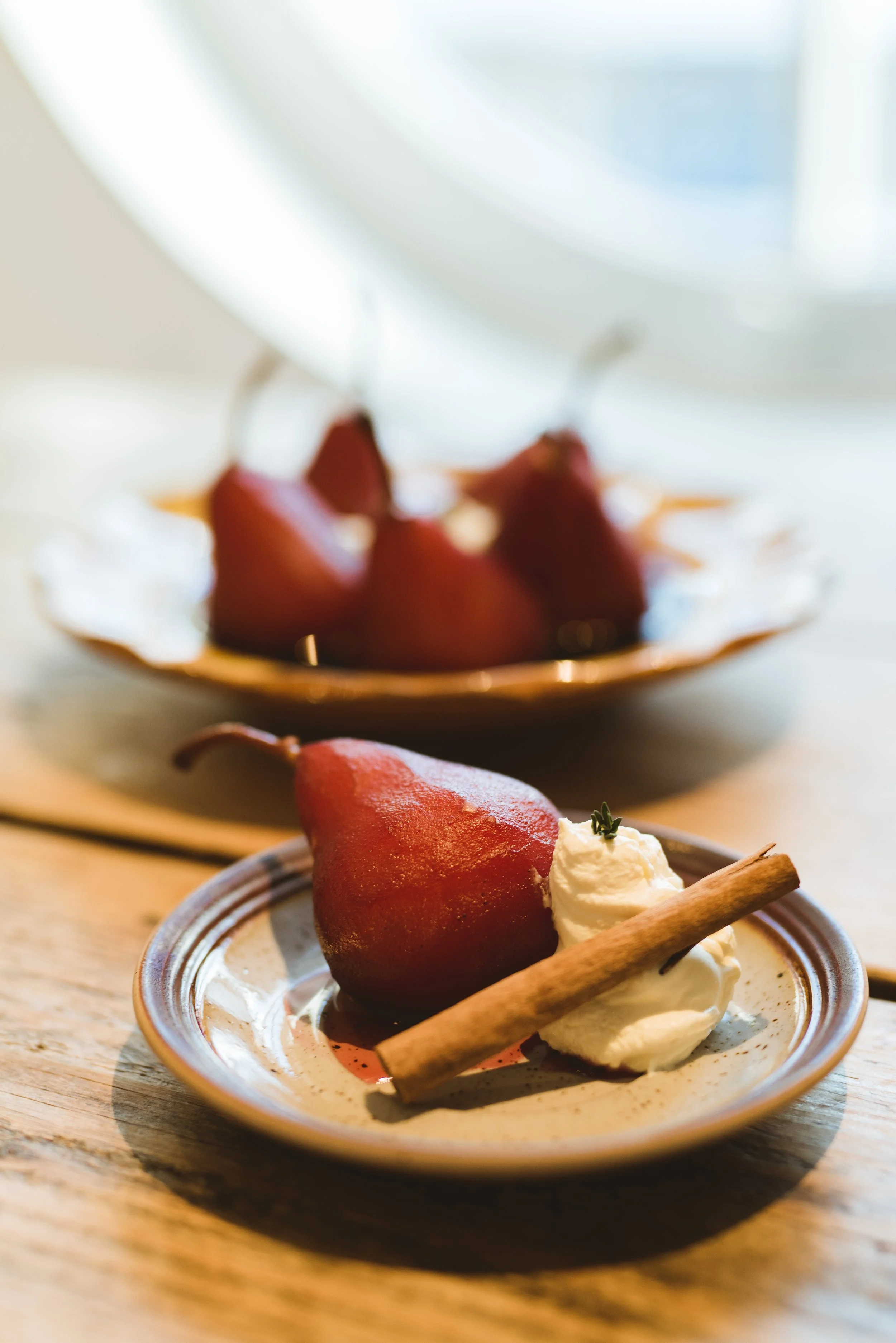 Close-up of a pear served with whipped cream, a cinnamon stick, and a small sprig of herbs on a ceramic plate; blurred plate of pears in the background.