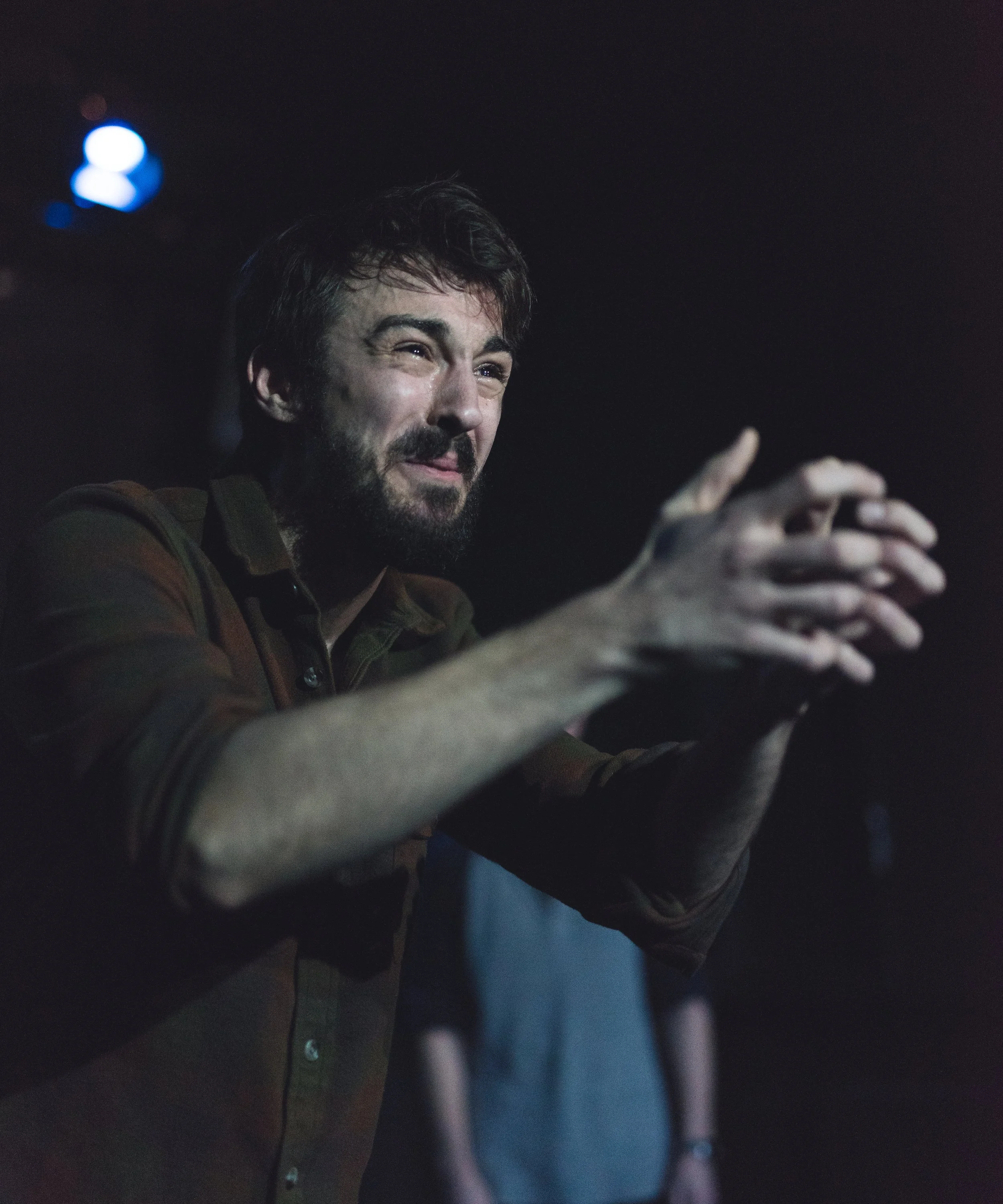 A man with dark hair and a beard, wearing a brown shirt, is smiling and gesturing with his hands in a dark setting with blue and pink lighting.
