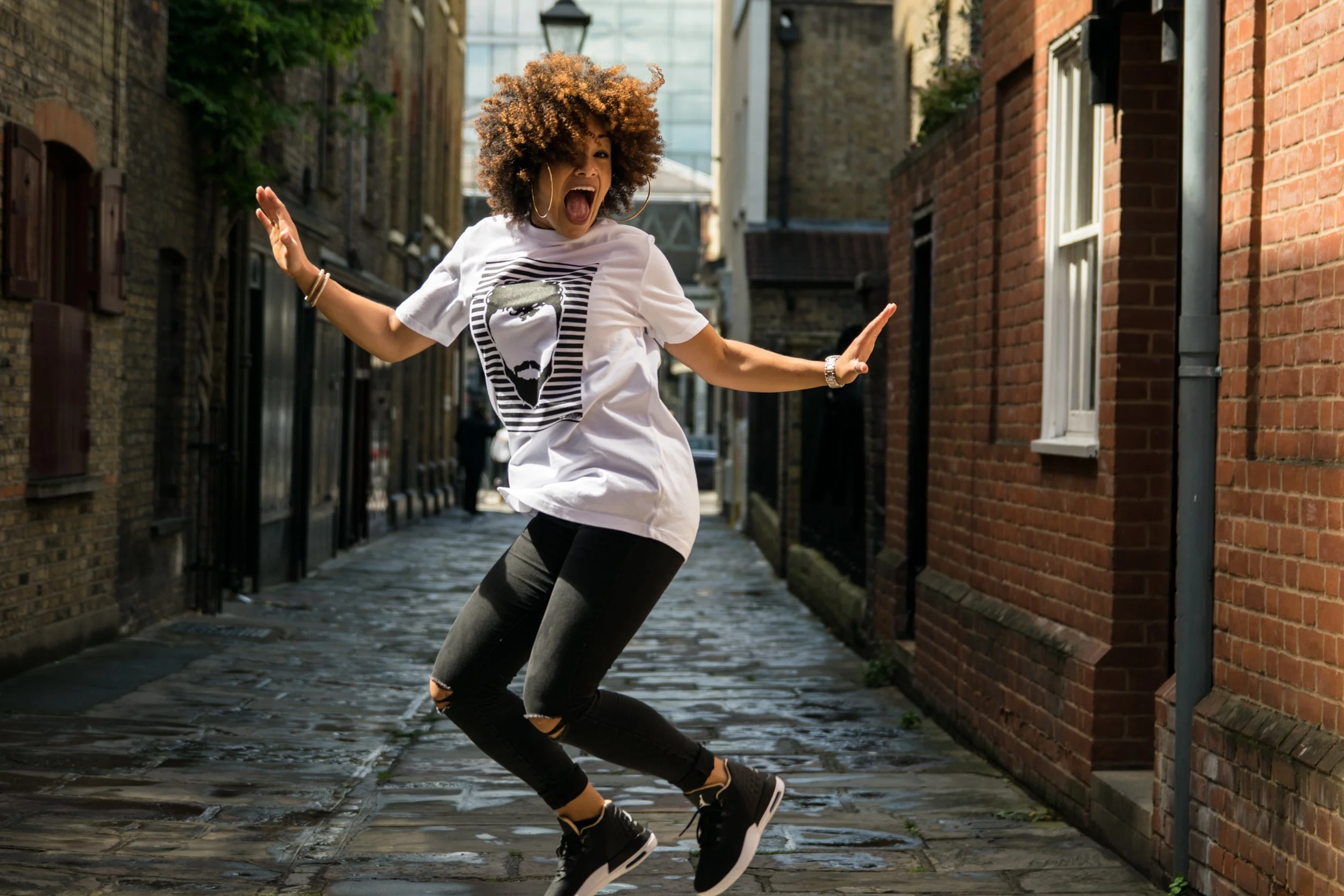 A woman with curly hair joyfully dancing on a cobblestone street between brick buildings.