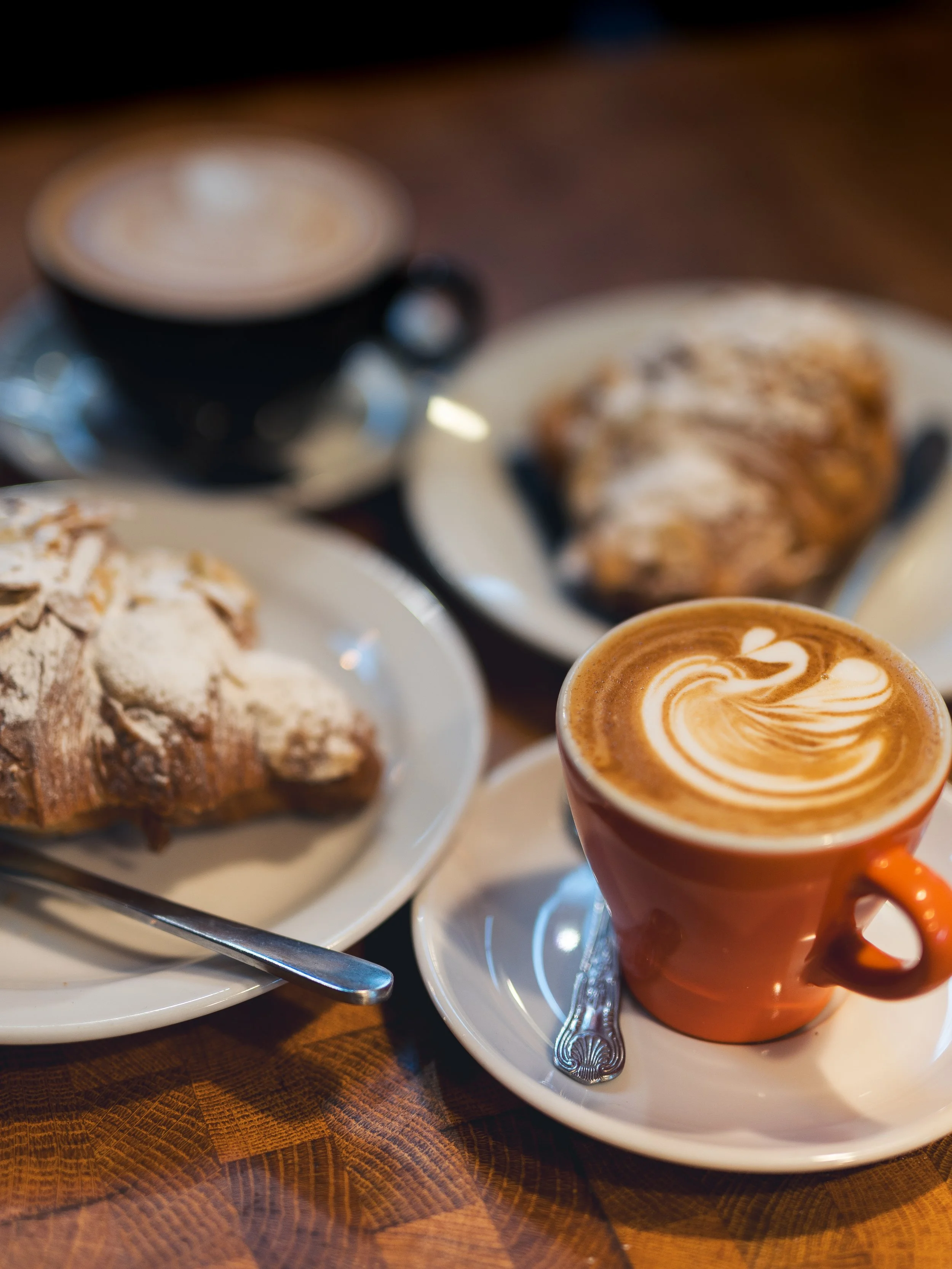 Close-up of a coffee cup with latte art, two plates of croissants dusted with powdered sugar, and a cup of coffee in the background on a wooden table.