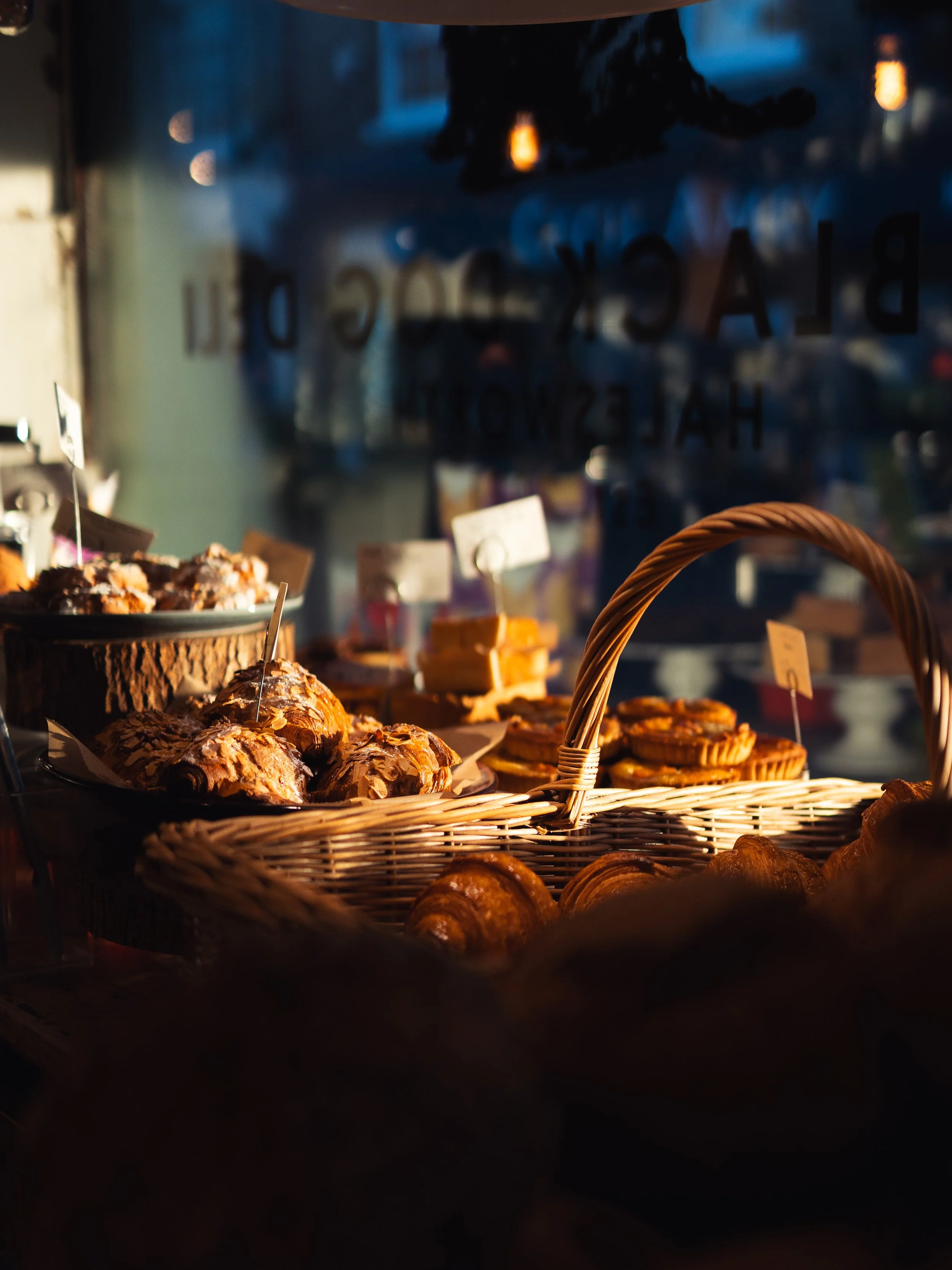 Bakery display with croissants and other pastries in warm sunlight, seen through a glass window.
