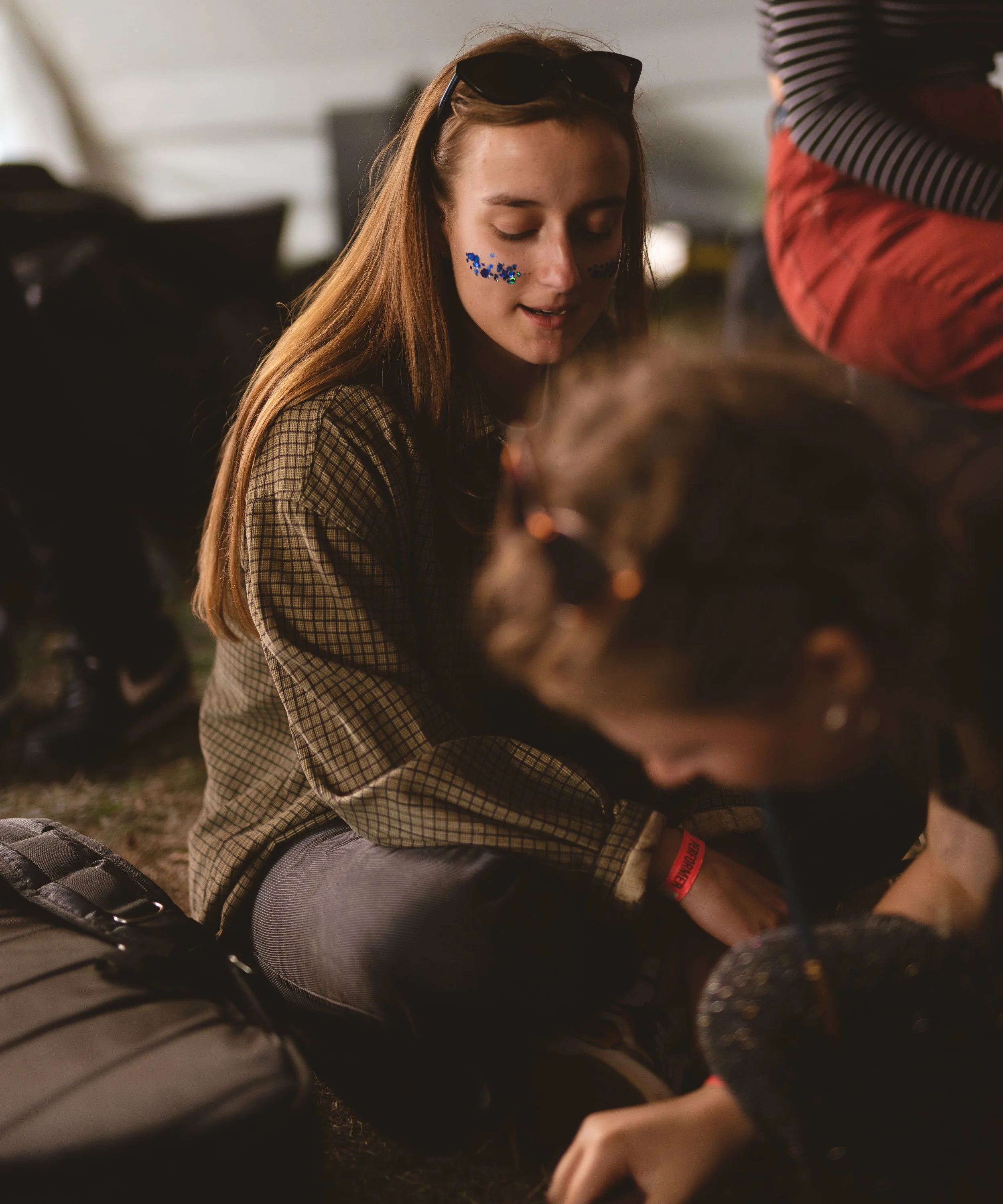 A young woman with long red hair, sunglasses on her head, and blue glitter face paint on her cheeks, sitting on the ground and smiling while talking to another person near her.