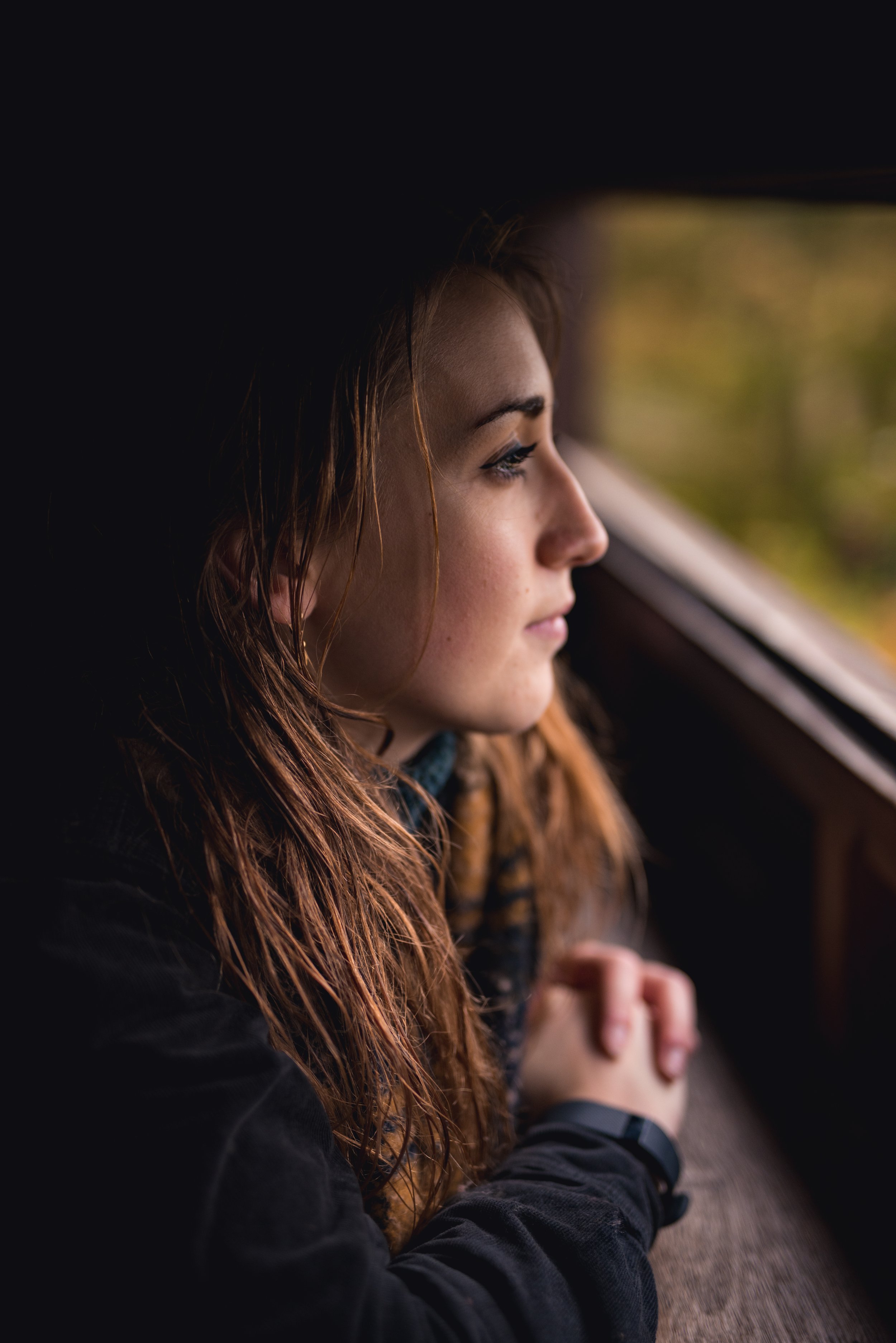 A young woman with wavy reddish hair looking out a window, appearing deep in thought.