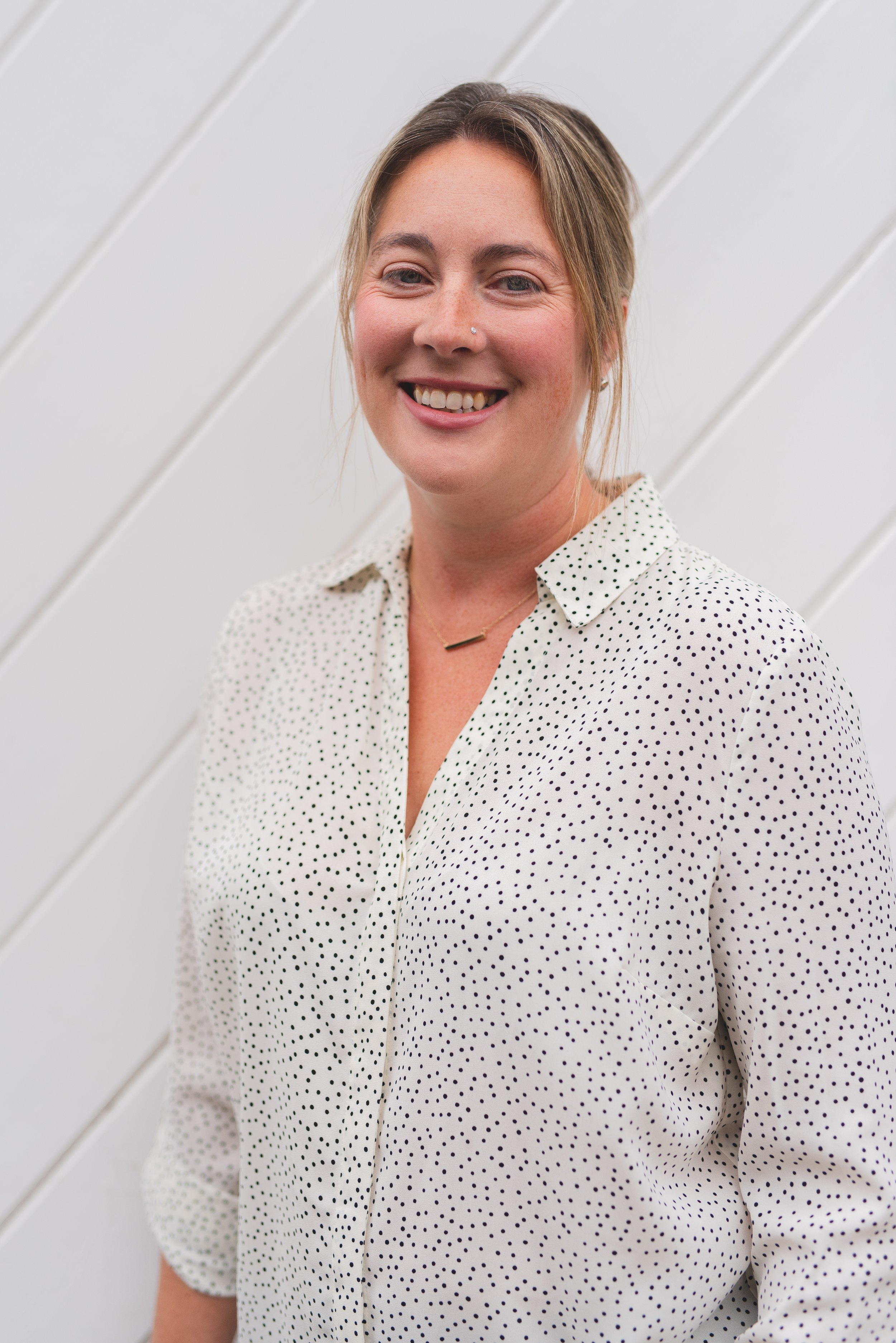 A smiling woman with blonde hair in a loose bun, wearing a white blouse with black polka dots and a small gold necklace, standing against a white paneled wall.