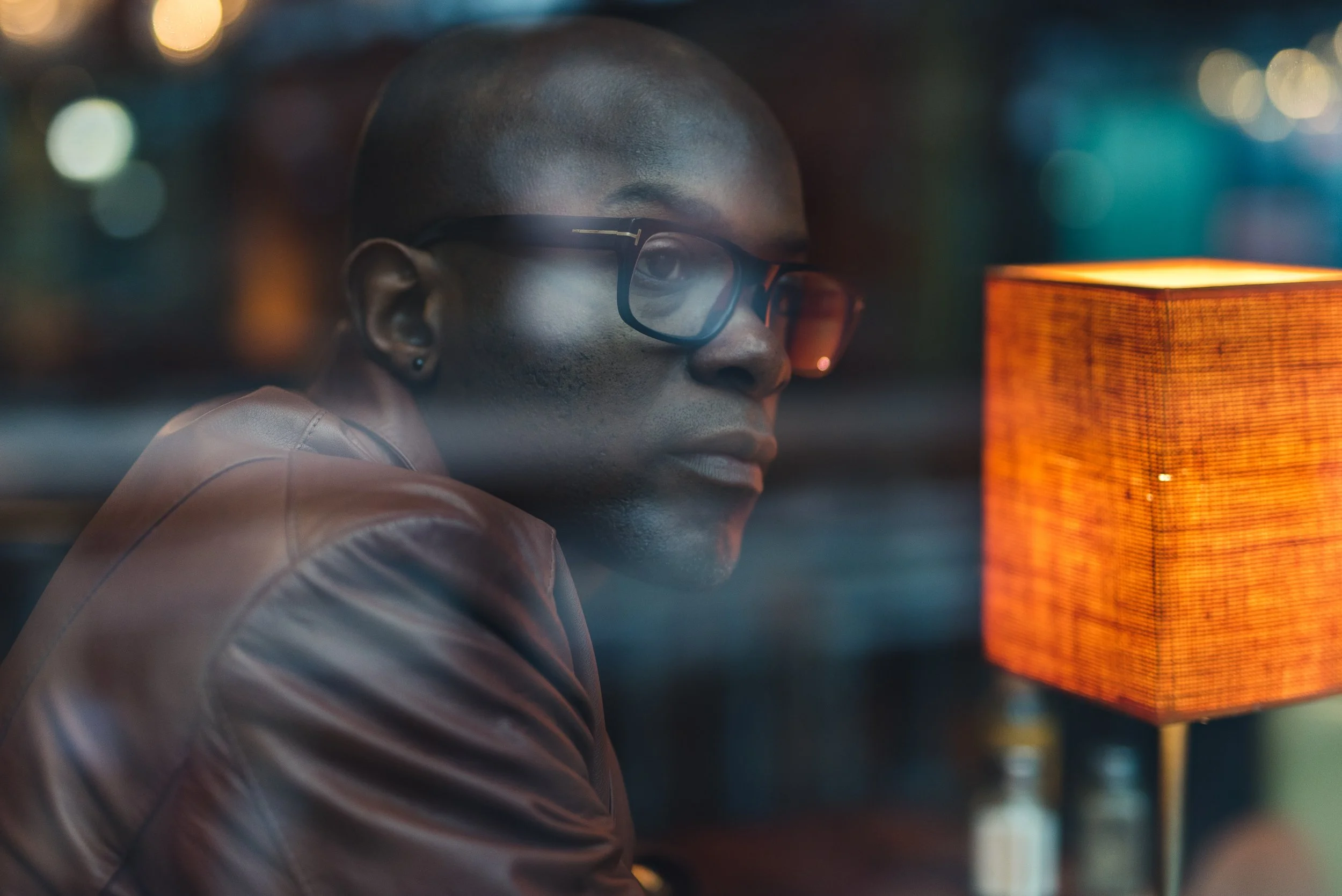A man with dark skin, short hair, wearing glasses and a brown leather jacket, sitting at a table with an orange lamp, looking out a window at night with city lights in the background.