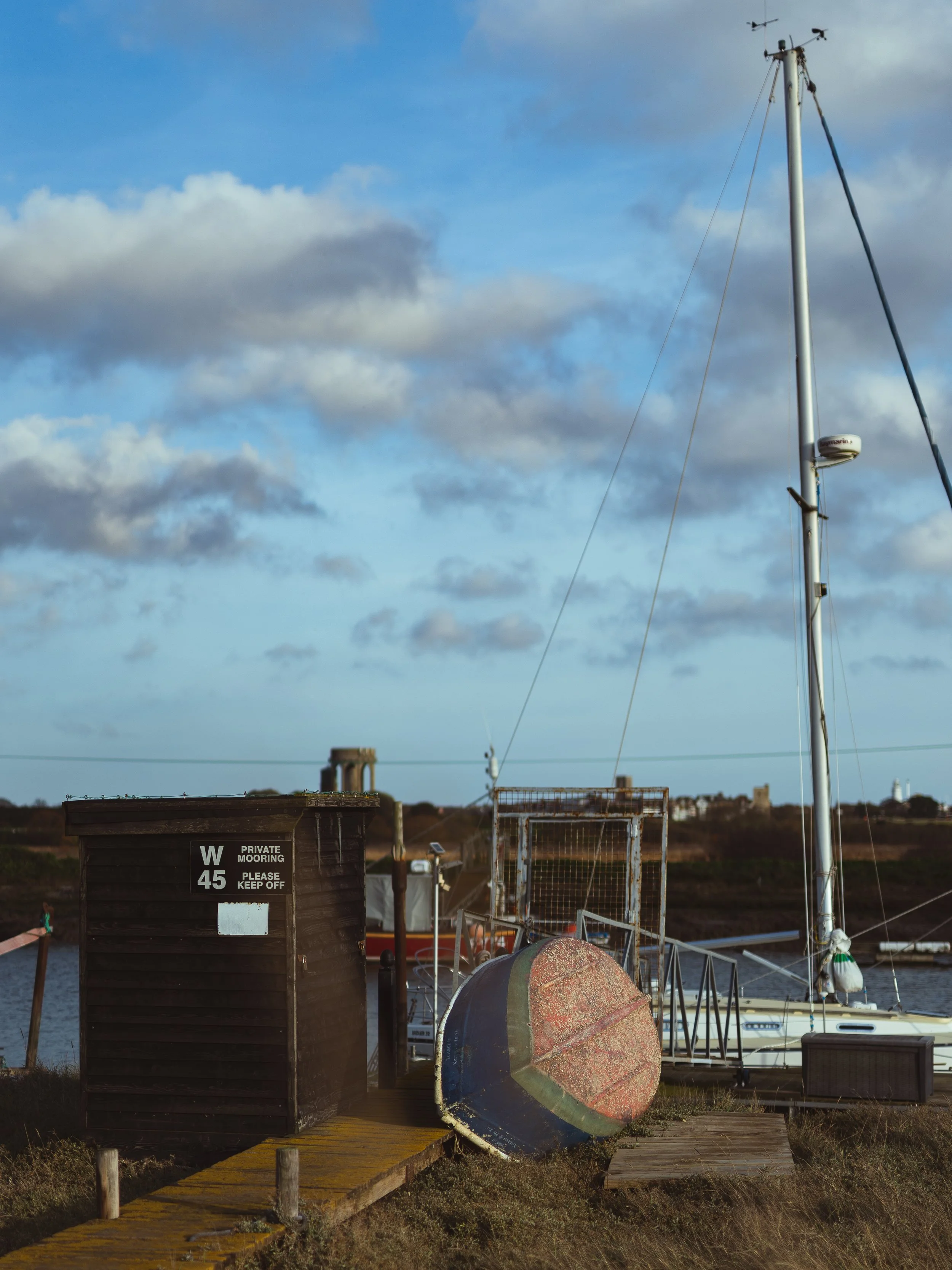 Sailboat docked beside a wooden shed, with a large, weathered rowboat on the ground, under cloudy blue sky.