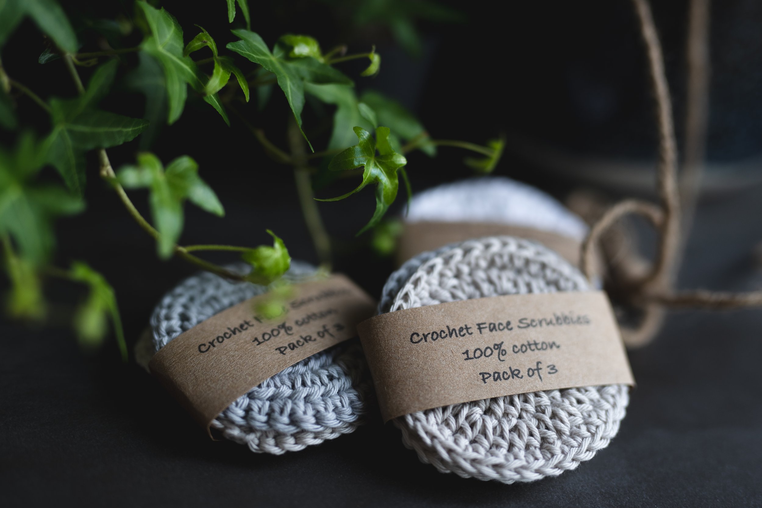 Three soap scrubbies wrapped in brown paper labels that read "Crochet Face Scrubbies, 100% cotton, Pack of 3" are placed on a black surface, with green ivy leaves nearby.