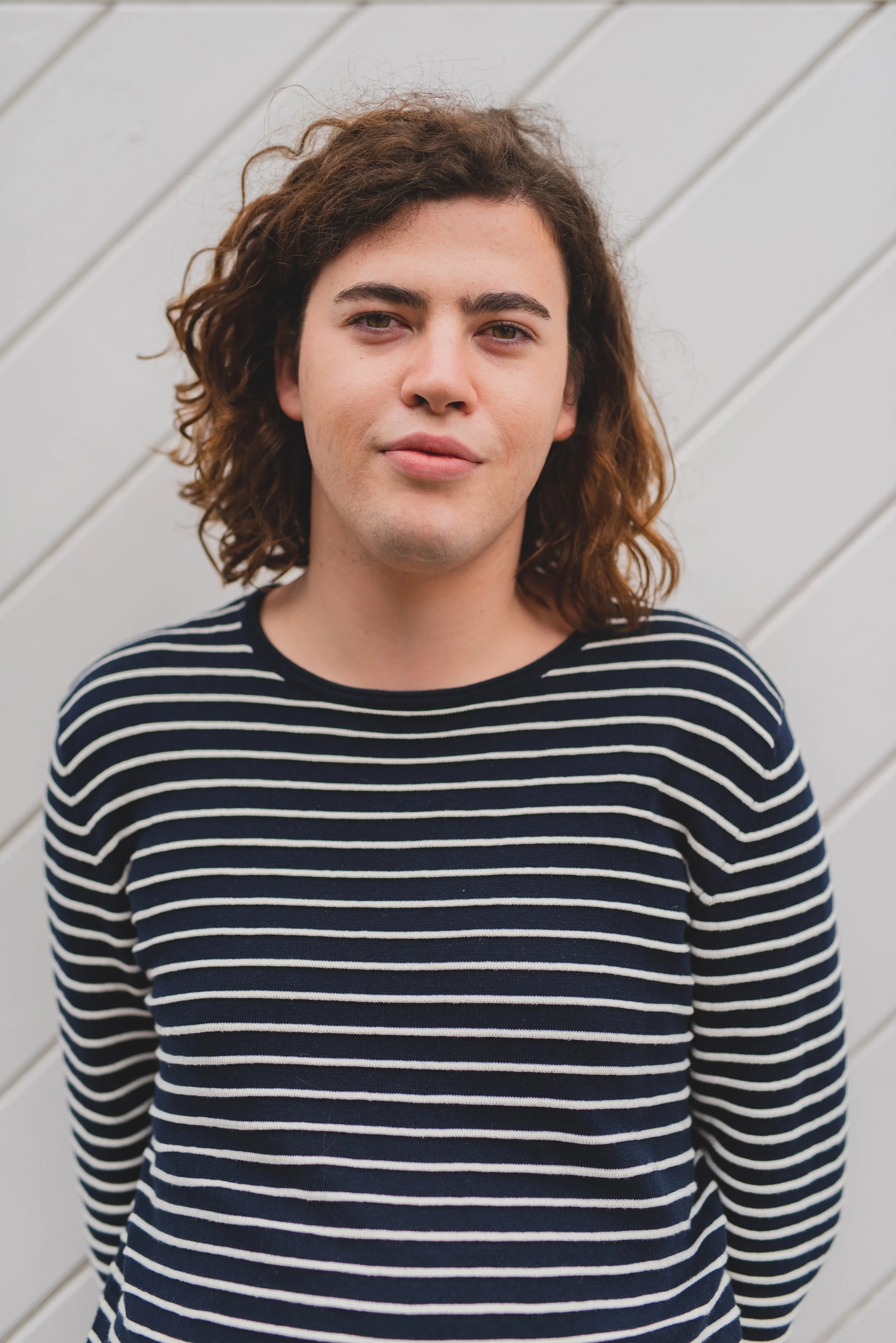 A young person with shoulder-length curly brown hair, wearing a black and white striped long-sleeve shirt, stands in front of a white wooden wall.