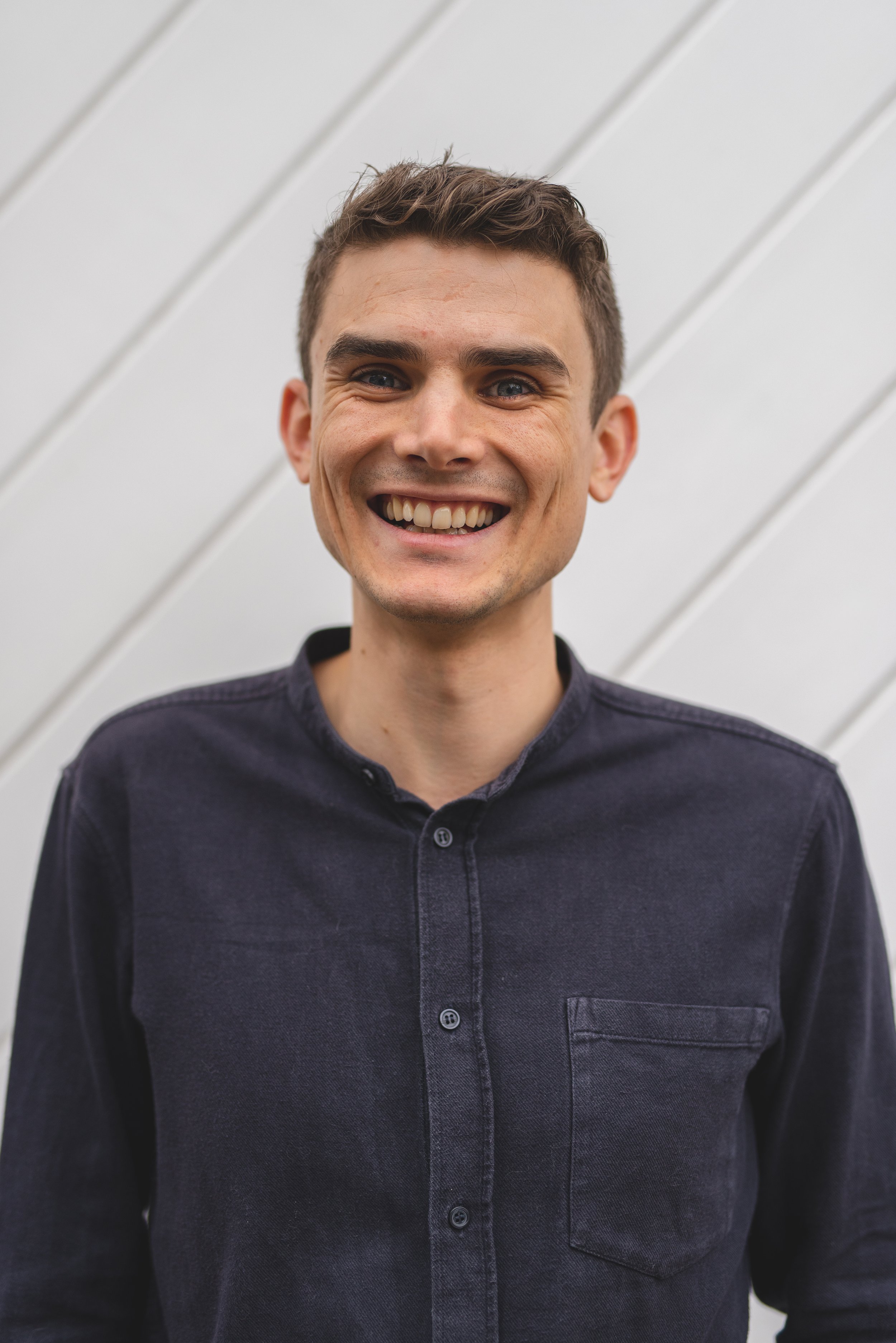 A young man with short brown hair and blue eyes smiling, wearing a dark button-up shirt, standing in front of a white wall with diagonal planks.