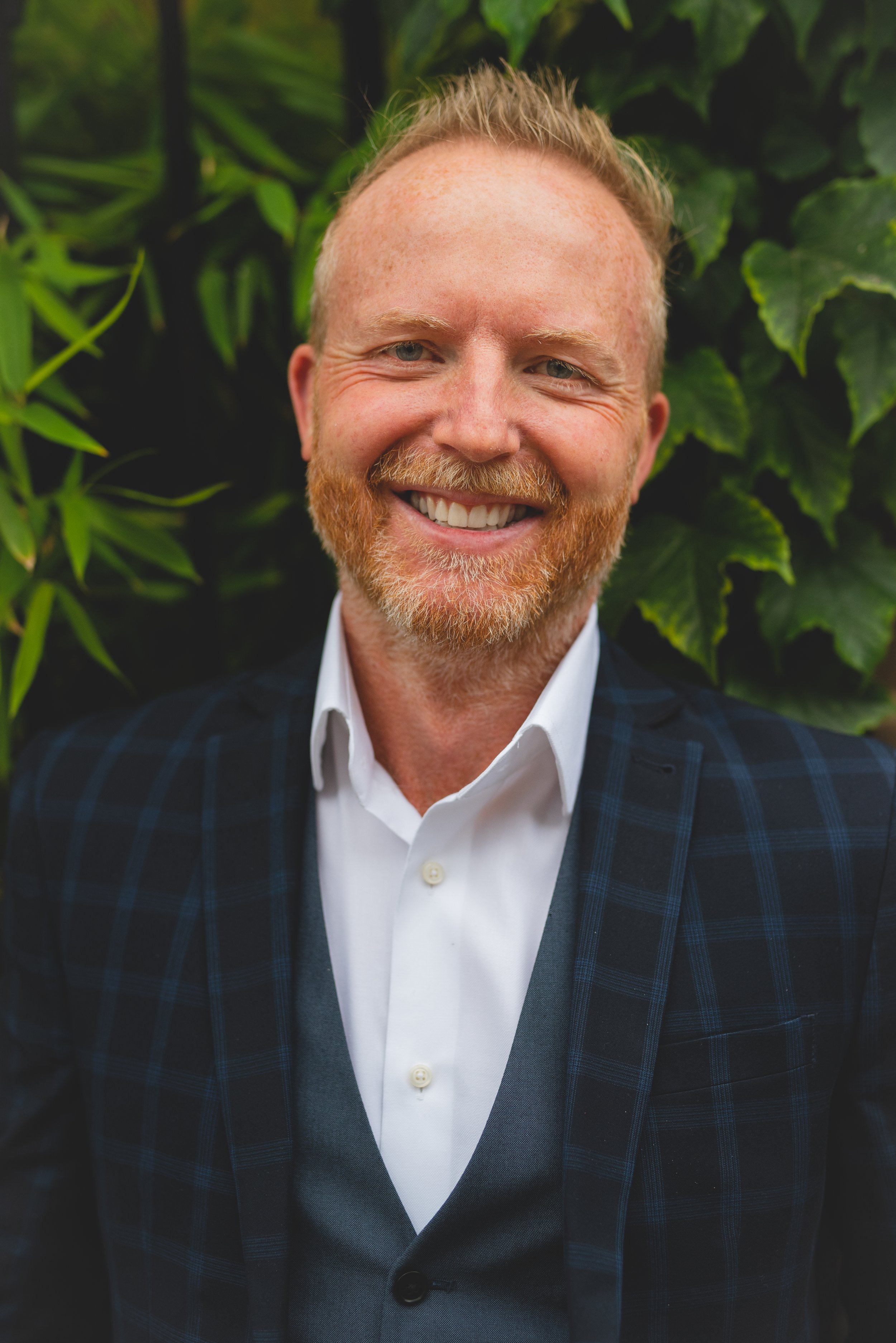 A smiling man with red hair and a beard, wearing a white shirt and a blue plaid blazer, standing outdoors in front of green foliage.