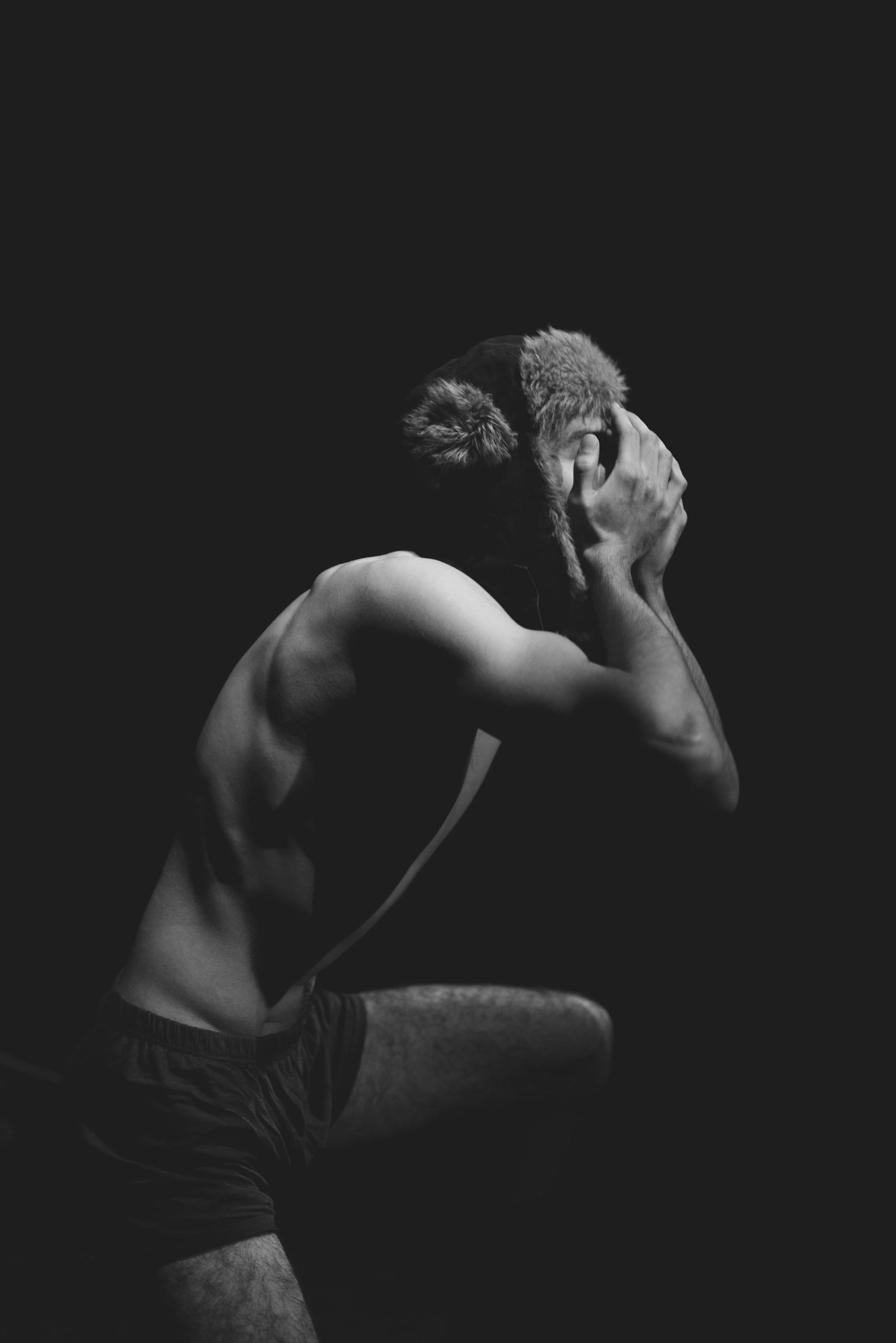 A black-and-white photo of a shirtless man with a beard, wearing a furry hat, covering his face with both hands, sitting against a dark background.