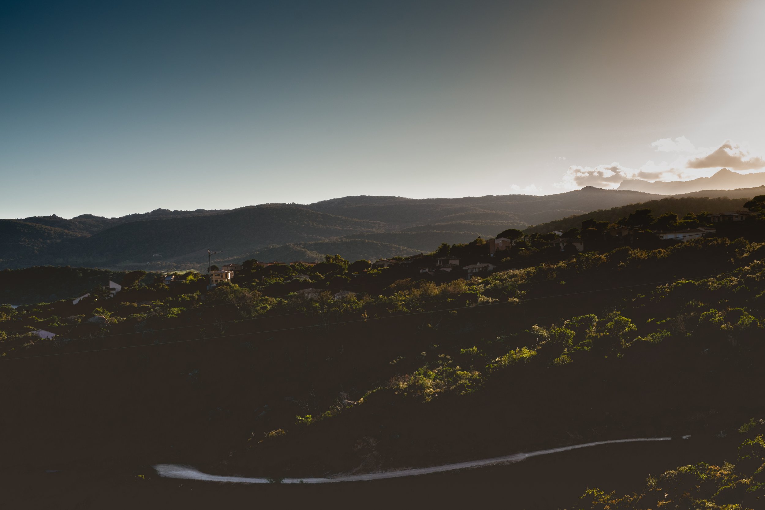 Landscape of rolling hills and mountains with scattered houses, under a sky with sunlight and some clouds.