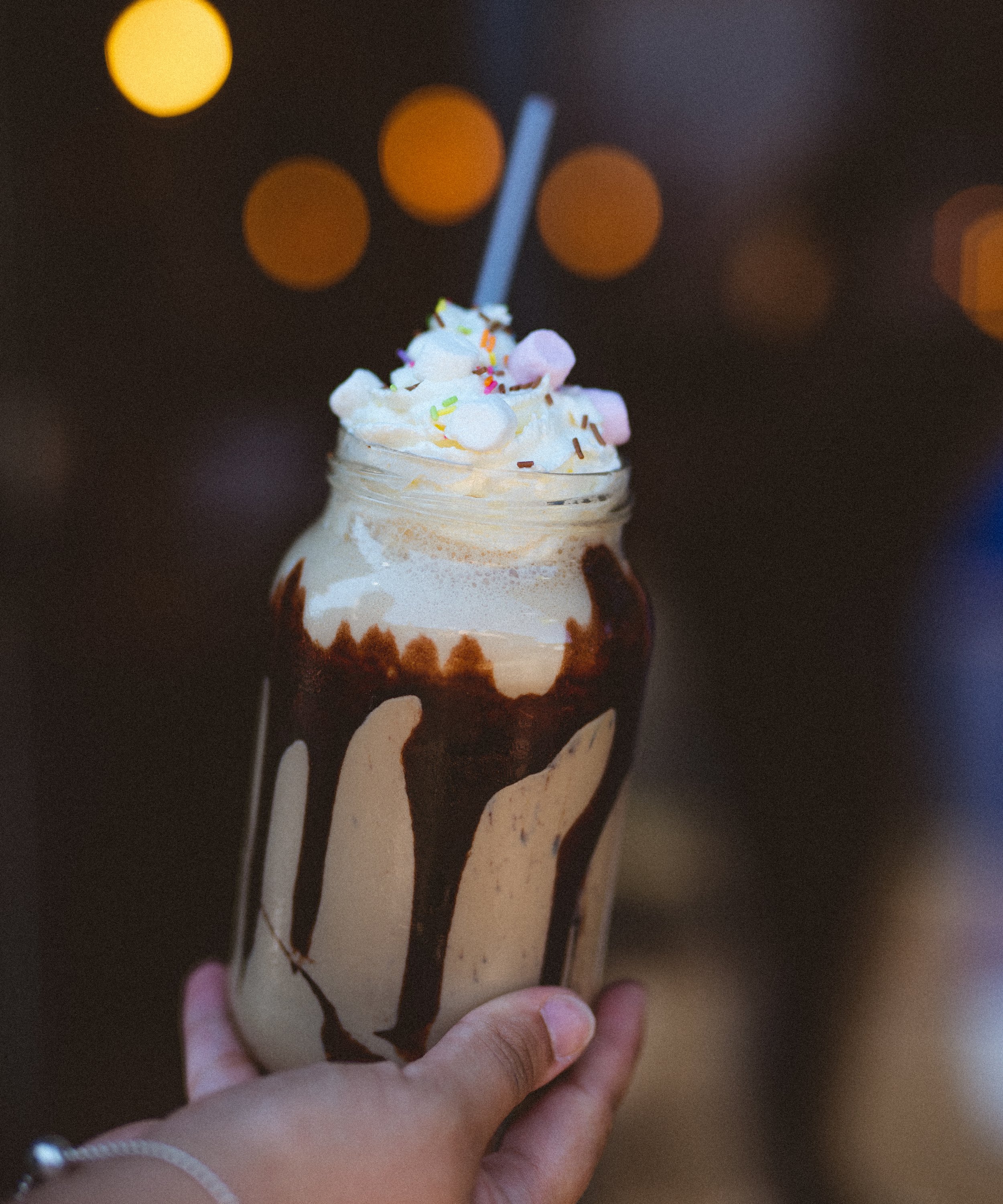 Person holding a milkshake topped with whipped cream, marshmallows, and rainbow sprinkles, with chocolate syrup dripping down the jar, against a blurred background with bokeh lights.
