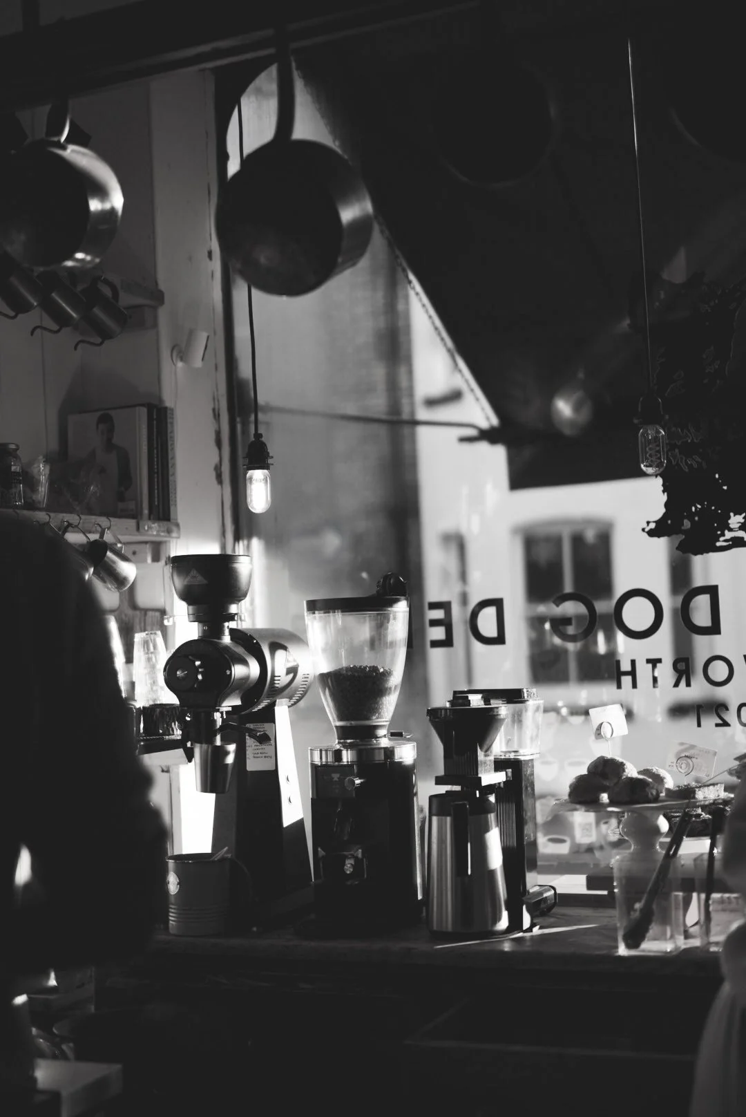 Black and white photo of a coffee shop interior with coffee brewing equipment, mugs, jars, and a window with text that says 'Doge De' and 'North' visible.
