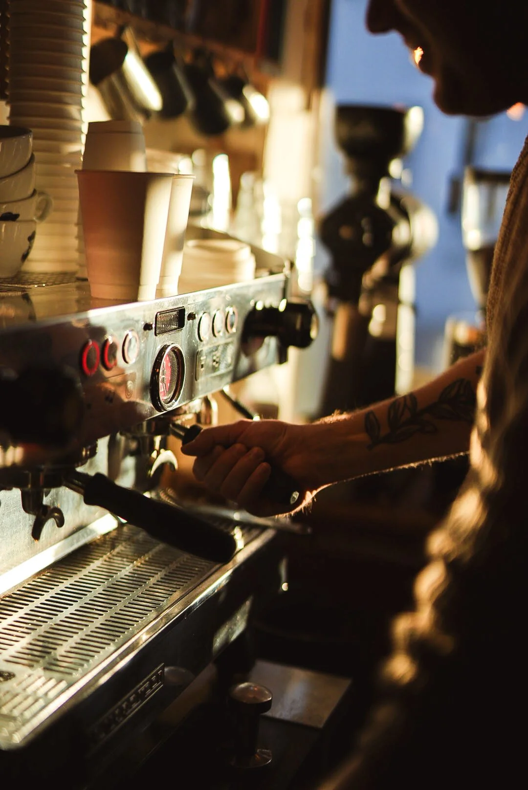 Barista preparing coffee on an espresso machine, with cups and mugs on shelves in a coffee shop.