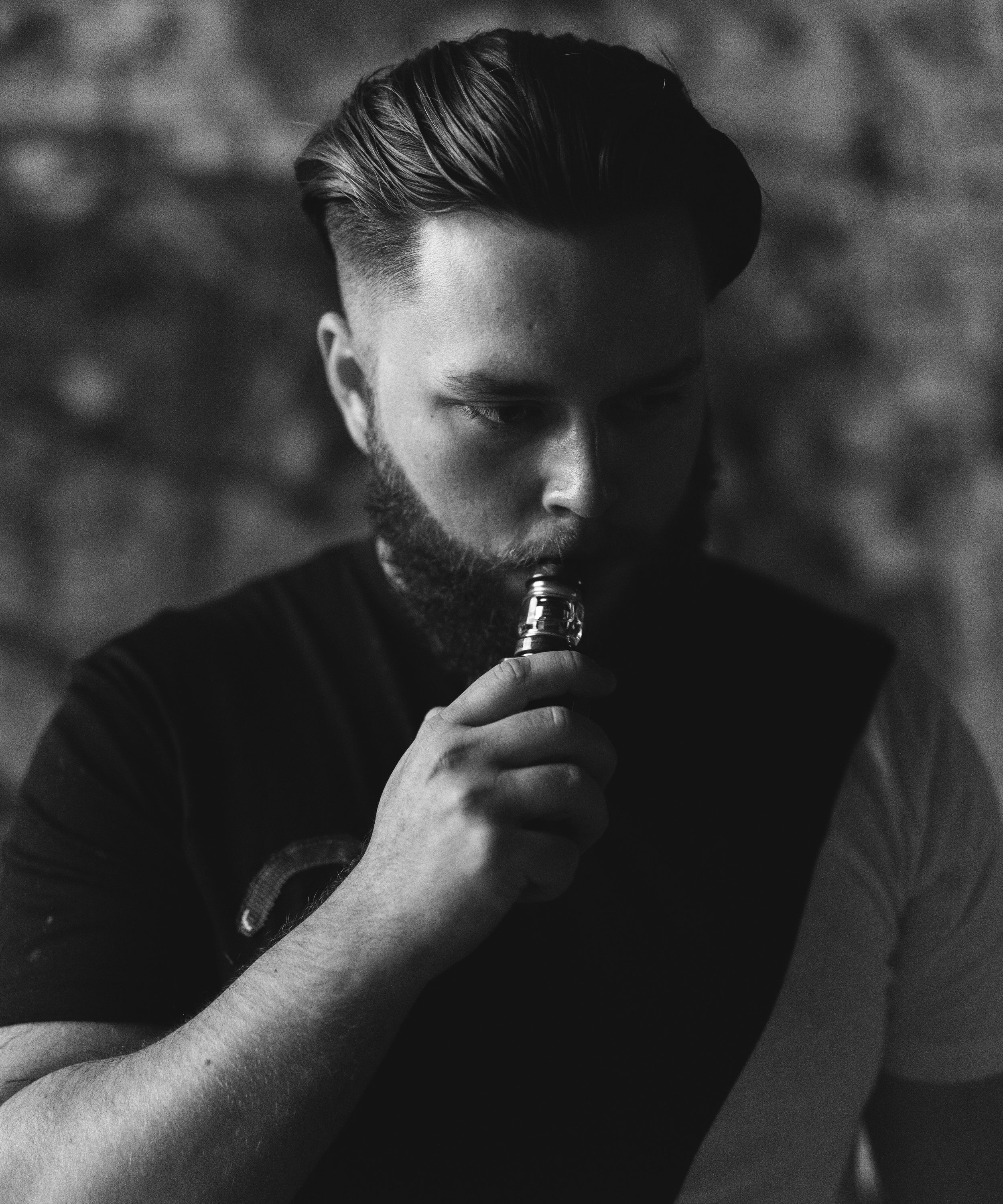 A young man with a styled haircut and beard, holding an e-cigarette near his mouth, in a dimly lit setting with a blurred brick wall background.