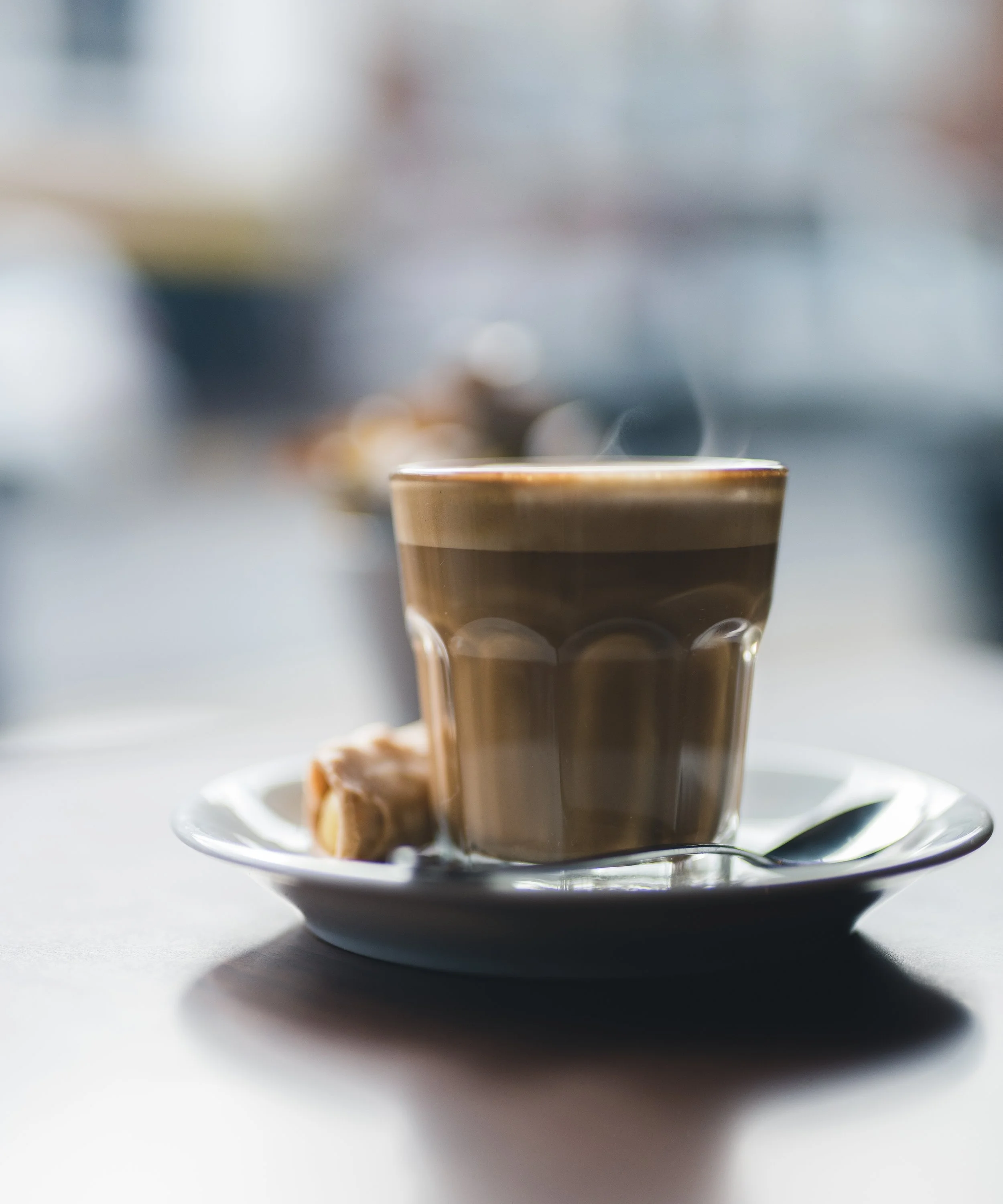 A steaming cup of coffee on a white saucer with a spoon and a small pastry on the side, on a bright table.